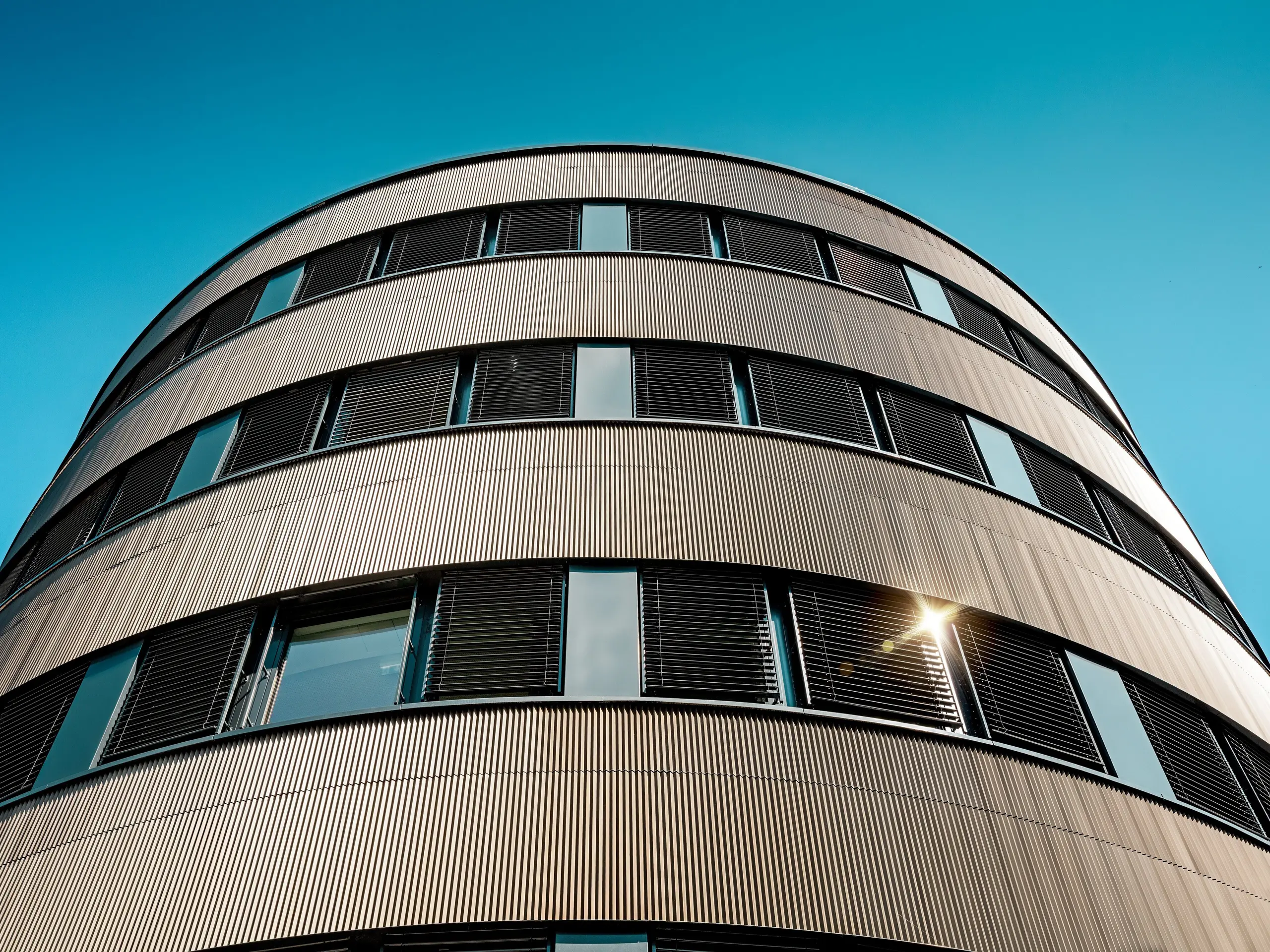 Vue spectaculaire depuis le bas du nouveau bâtiment incurvé du commissariat de police 43 à Hambourg : la façade en aluminium PREFA, composée de profils extrudés anodisés « triangle » couleur bronze moyen, révèle tout son potentiel et attire immédiatement le regard. Sa structure finement nervurée reflète la lumière du soleil dans des nuances vibrantes et souligne la précision des courbes et les bandes horizontales des fenêtres. L’aluminium PREFA séduit par sa haute résistance aux intempéries, sa protection contre la corrosion, sa durabilité et ses exigences minimales d’entretien – une solution idéale pour des façades durables et architecturalement sophistiquées.