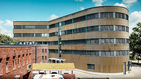 Vue extérieure dynamique du commissariat de police 43 agrandi à Hambourg : le nouveau bâtiment incurvé, revêtu d’environ 1 100 m² de profils extrudés en aluminium PREFA anodisé bronze moyen « triangle », crée un contraste saisissant avec la maçonnerie existante. La surface finement texturée joue avec la lumière et l’ombre, conférant à la façade profondeur et précision. Grâce à leur haute résistance à la corrosion, leur durabilité et leur faible besoin d’entretien, les solutions en aluminium PREFA sont synonymes de construction durable, fonctionnelle et d’une grande sophistication architecturale.