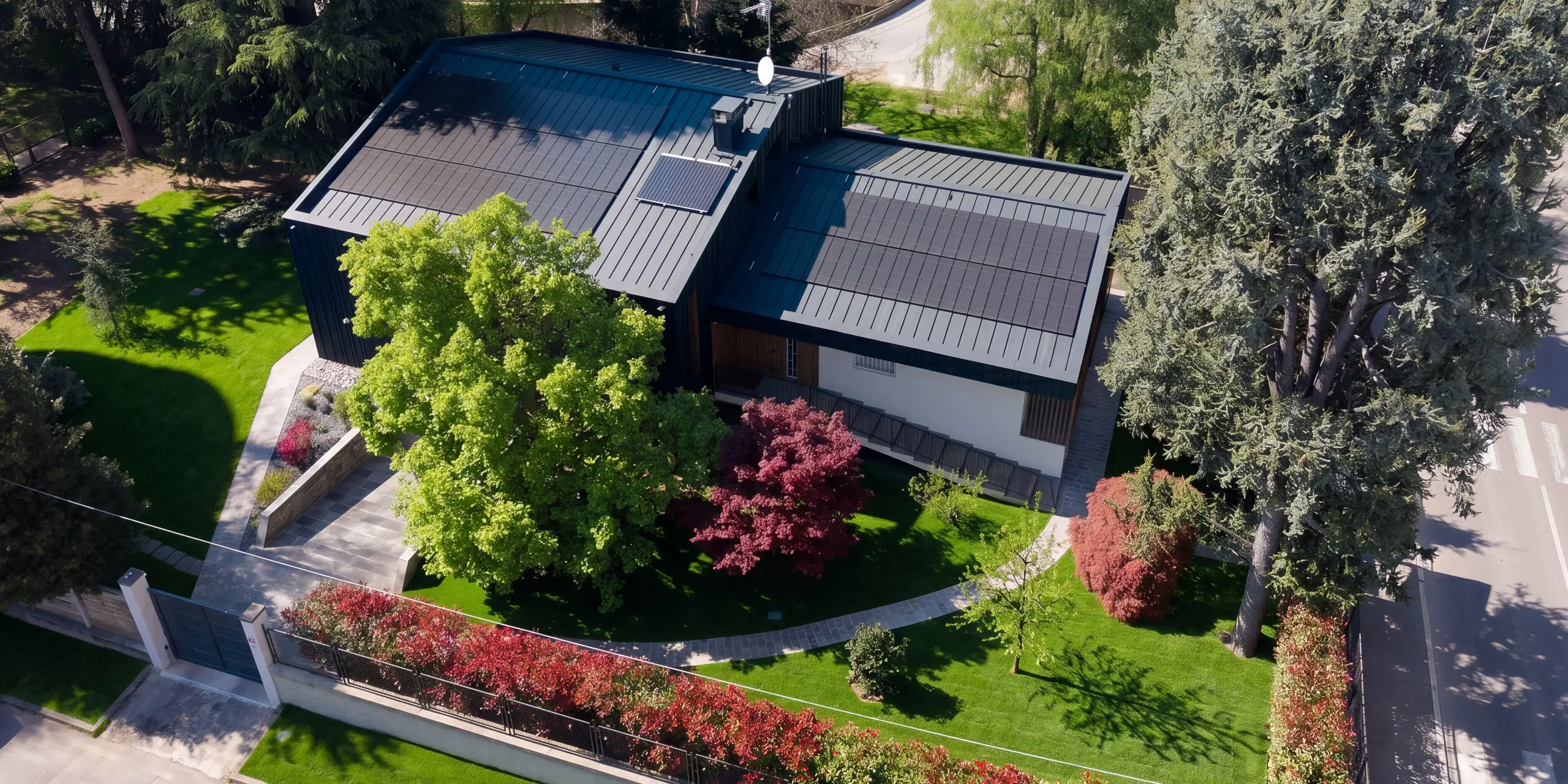Vue aérienne spectaculaire de la Villa Castellanza, dotée d'une toiture et d'une façade PREFALZ couleur P.10 Anthracite. De grands modules solaires PREFALZ intégrés définissent l'enveloppe moderne du bâtiment et permettent une production d'énergie durable. Nichée dans un jardin soigné et arboré, la résidence illustre l'alliance parfaite entre l'aluminium PREFALZ, le design et la fonctionnalité.