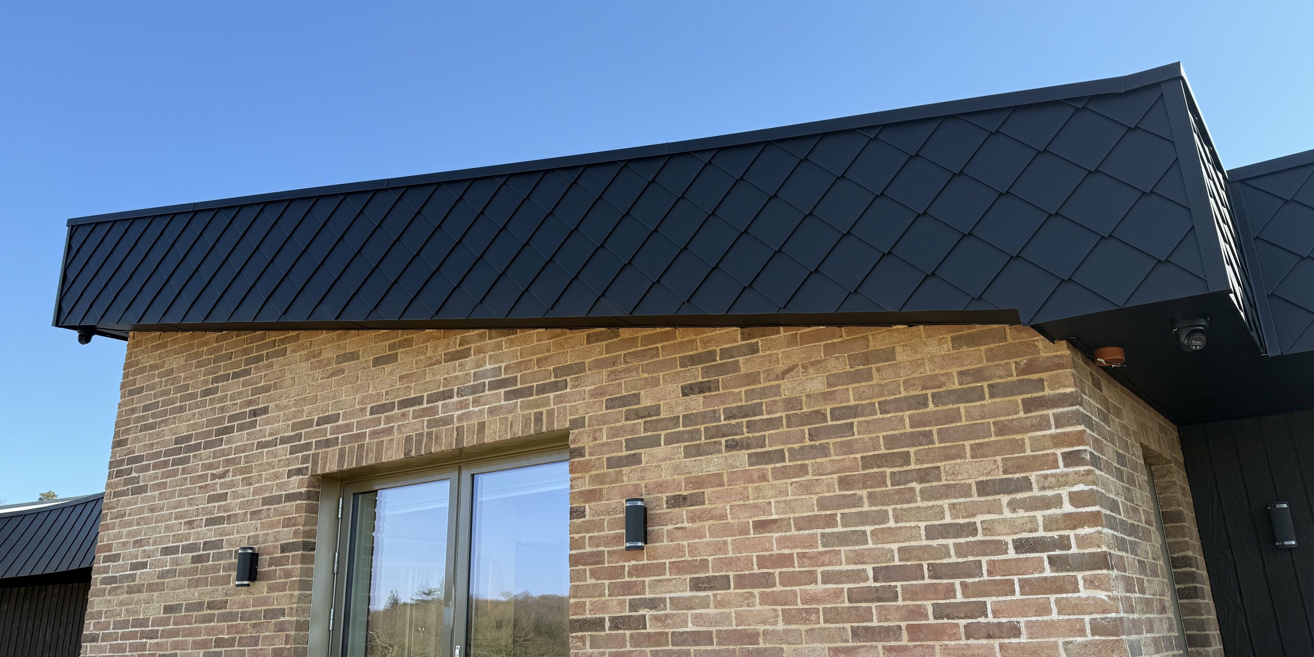 Maison individuelle moderne à la façade remarquable : l’alliance de briques de parement chaleureuses et d’élégants losanges de façade PREFA 20 x 20 couleur anthracite P.10 confère à la façade un fort impact visuel. Les robustes bardeaux métalliques apportent à la maison un design exclusif et impressionnent par leur durabilité, leur résistance aux intempéries et leur facilité d’entretien. Sur un ciel d’un bleu éclatant, la structure géométrique se détache particulièrement et souligne la grande qualité architecturale.