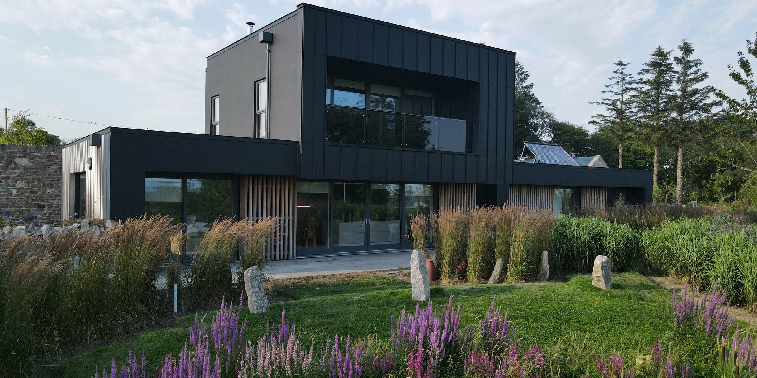 L'image présente une maison d'architecte moderne à Wexford, entourée d'un jardin luxuriant aux herbes hautes et à la lavande en fleurs violettes. La façade noire PREFALZ, coloris P.10 noir, confère à la maison une allure saisissante grâce à ses lignes épurées, sa finition noir mat profond et sa structure verticale précise. Les larges baies vitrées et le balcon couvert soulignent son caractère moderne. Robuste, la façade en aluminium PREFA est résistante aux intempéries et durable. Elle crée un contraste saisissant avec la végétation luxuriante au premier plan.