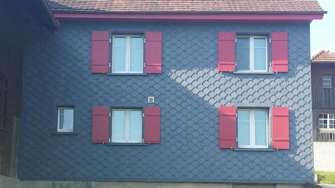 Rénovation de façade à l’aide de losanges de façade PREFA, avec quatre fenêtres dotées de volets rouges