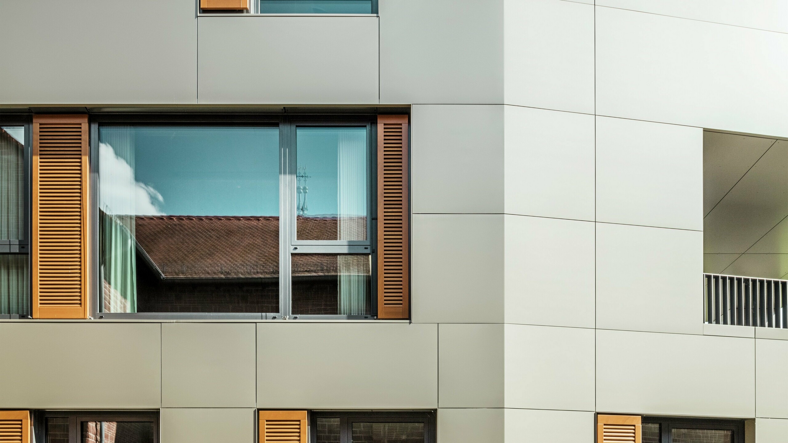 Détail de la façade de la mairie d'Alzenau, en Allemagne, avec des panneaux composites en aluminium PREFA de couleur bronze. Les grandes fenêtres sont encadrées d'élégants stores vénitiens en bois brun chaud qui confèrent à la façade une texture attrayante. Au premier plan, on aperçoit un parterre de fleurs soigné qui complète le design urbain du bâtiment avec des accents verts. Le ciel clair et les lignes épurées de l'architecture soulignent le caractère moderne du bâtiment.