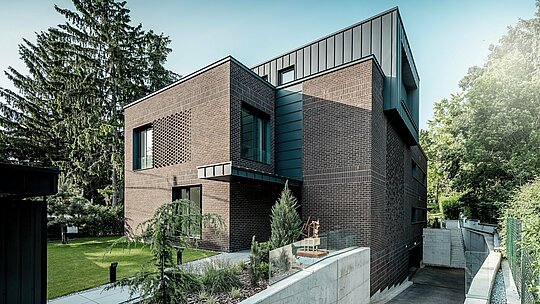 La photographie met en valeur l'architecture moderne de cette villa multigénérationnelle de Budapest, caractérisée par une remarquable combinaison de briques rouges foncées et d'aluminium PREFALZ P.10 anthracite. Le cube aux contours nets, avec ses joints verticaux, contraste fortement avec la façade en briques texturées. L'élégante allée menant au garage souterrain et le jardin paysager soigné soulignent la qualité de la construction. Les produits en aluminium PREFA, durables et résistants aux intempéries, confèrent au bâtiment une allure intemporelle, précise et d'une grande expressivité architecturale.