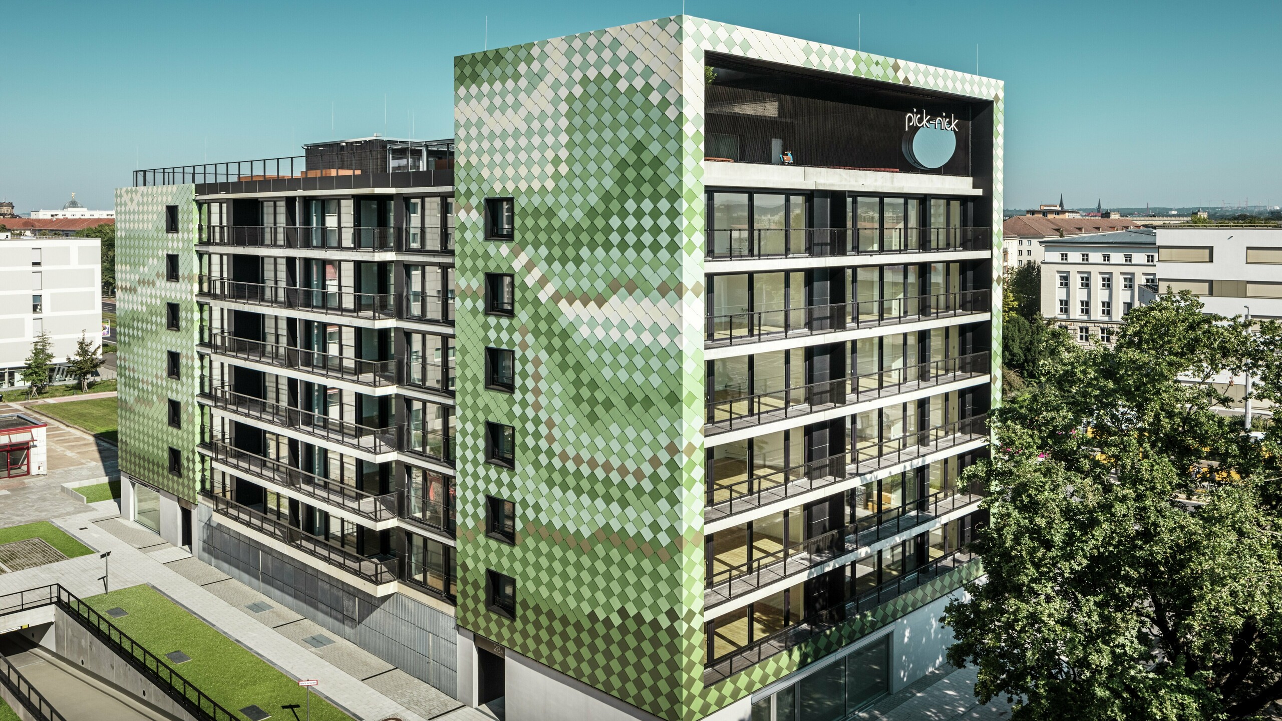 Bâtiment résidentiel et commercial "Pick-Nick" avec une façade PREFA frappante en losanges d'aluminium dans divers tons verts et beiges. Le concept de couleurs dynamique et la conception des lignes claires de la façade en aluminium sont en harmonie avec les grandes surfaces vitrées et les espaces verts environnants. L'inscription « Pick-Nick » sur le bord du toit souligne l'importance du bâtiment pour la ville de Dresde.