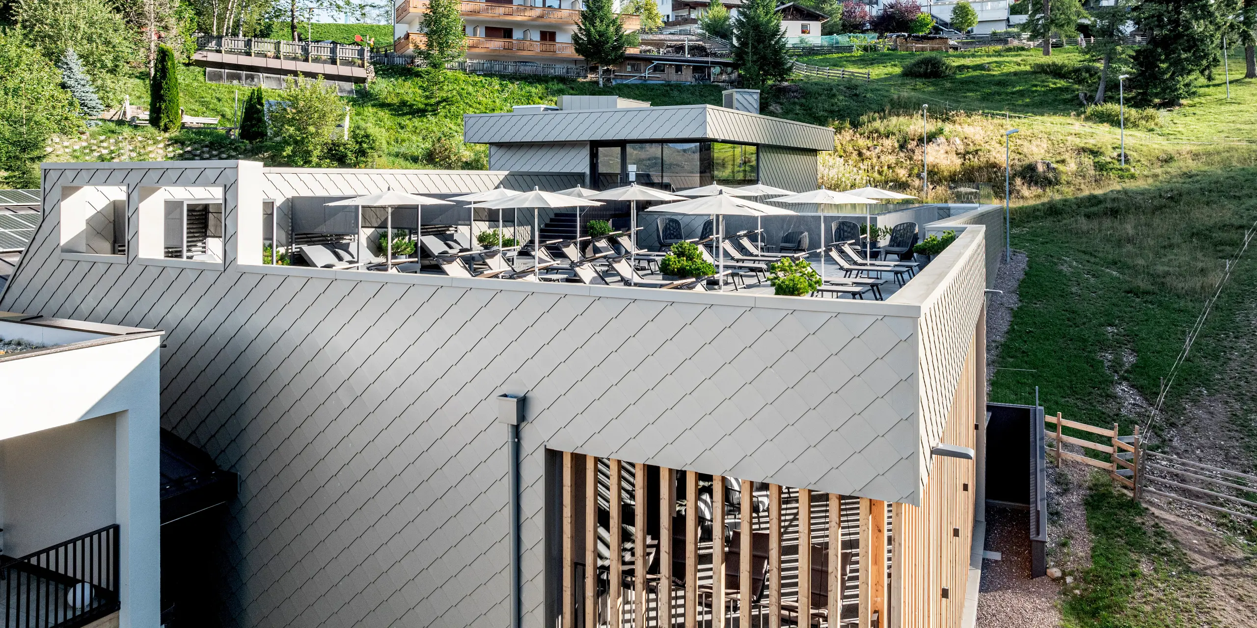 Terrasse surélevée du Sporthotel Obereggen, dans le Tyrol du Sud, avec piscine, espace détente, parasols et vue imprenable sur les pistes alpines et les chalets environnants. Les lignes épurées du bâtiment sont soulignées par la façade en aluminium PREFA, composée de losanges de façade 29x29 couleur bronze, qui confère à l'architecture une structure précise et dynamique. La teinte chaude du bronze s'harmonise avec le bois et la nature. Durables, résistants aux intempéries et nécessitant peu d'entretien, les produits en aluminium PREFA sont parfaitement adaptés à l'architecture hôtelière moderne en régions alpines.