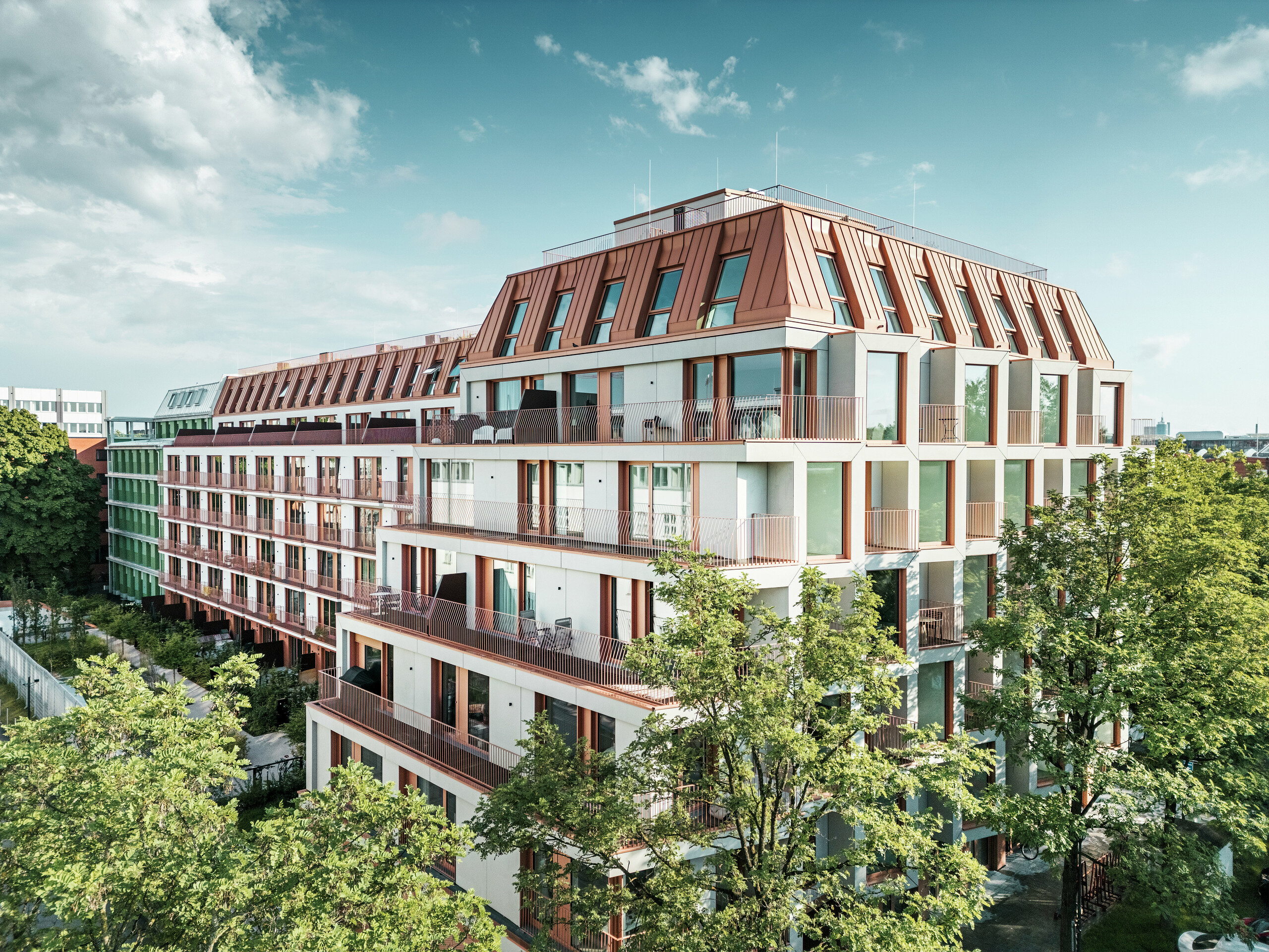 Vue d'ensemble du projet résidentiel urbain Van B à Munich avec une architecture claire, des espaces extérieurs verts et le toit en aluminium PREFALZ frappant en IGP Orange HDP de PREFA. Le matériau de toiture durable et nécessitant peu d'entretien constitue l'élément de conception unificateur du bâtiment et garantit une solution esthétique durable dans l'espace urbain. Des fenêtres généreuses, des balcons et des plans d'étage bien pensés offrent un confort de vie moderne avec une qualité de conception élevée.