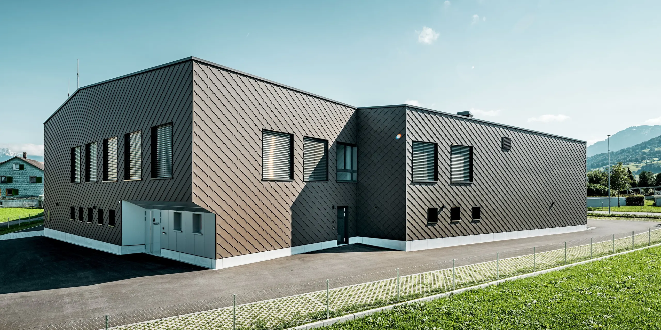 Vue arrière du bâtiment d'infrastructures abritant la caserne de pompiers et la centrale électrique de Wangen, en Suisse. Sa façade est revêtue en continu de losanges PREFA 29 × 29, coloris P.10 Brun. La structure losangique précise épouse la forme cubique du bâtiment, lui conférant une allure moderne et haut de gamme. La façade en aluminium durable séduit par sa grande résistance aux intempéries, ses faibles besoins d'entretien et la liberté de conception maximale qu'elle offre pour une architecture sophistiquée.