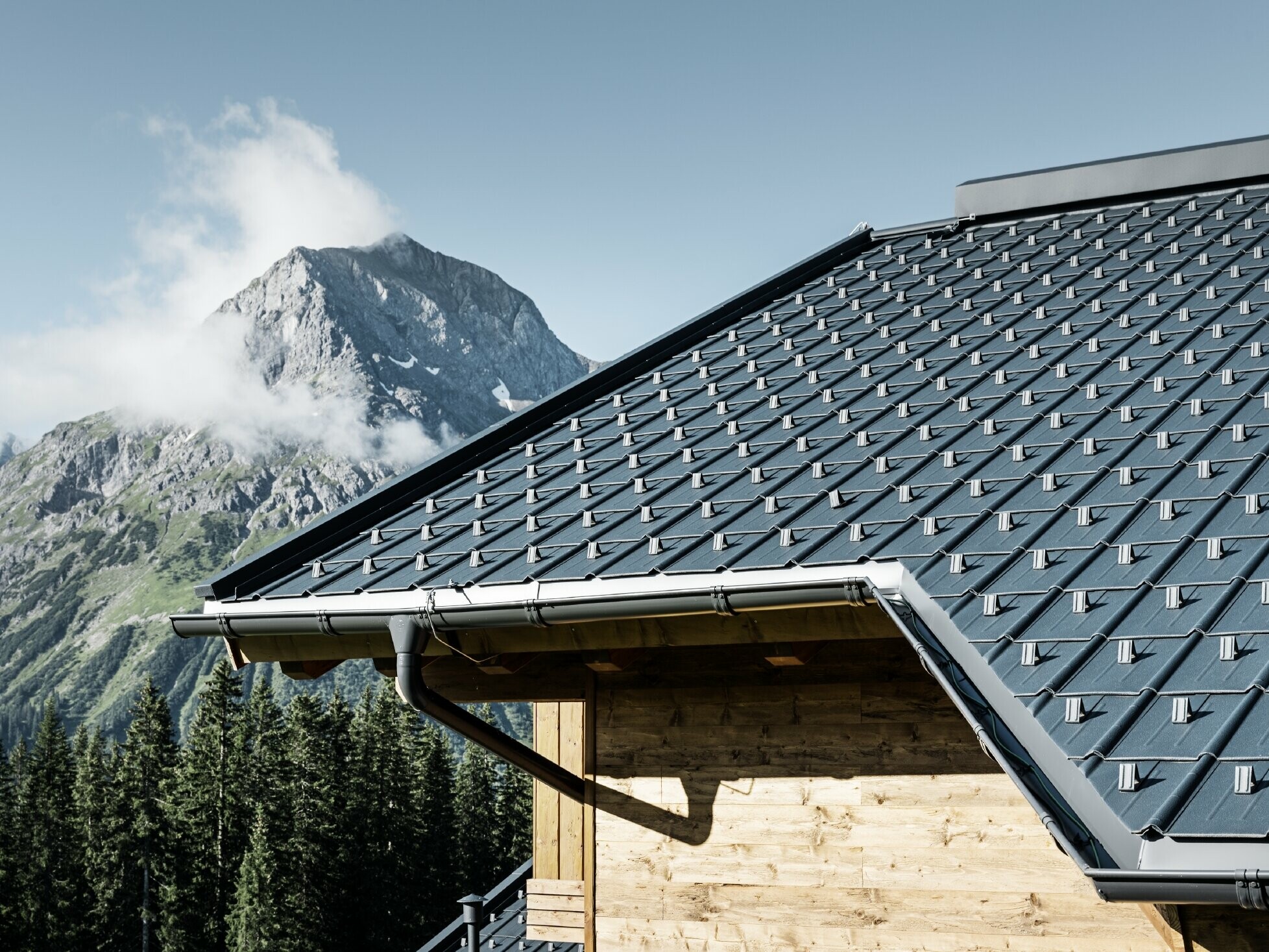 Tuile PREFA employée en région montagneuse - cabane recouverte avec des tuiles anthracite à Lech am Arlberg avec montagnes en arrière-plan ; gouttière PREFA anthracite ; façade en bois
