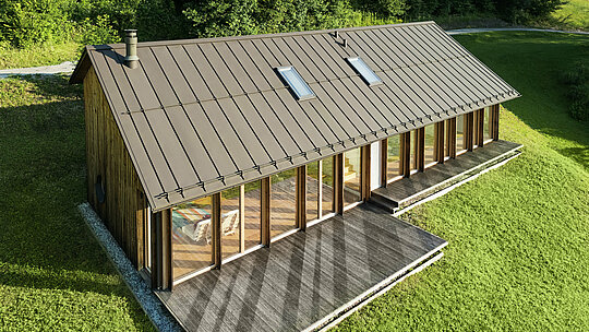 Maison en bois moderne avec une grande façade vitrée et un toit en aluminium PREFALZ résistant de couleur P.10 brun – idéale pour les terrains en pente comme à Vinje. La couverture de toit de haute qualité offre une résistance maximale aux intempéries, une longue durée de vie et une esthétique élégante. Grâce à la pose précise des joints debout et à l’intégration de fenêtres de toit, un concept d’habitat durable et baigné de lumière voit le jour. La vue en plongée met en évidence la finition soignée des joints debout en aluminium.