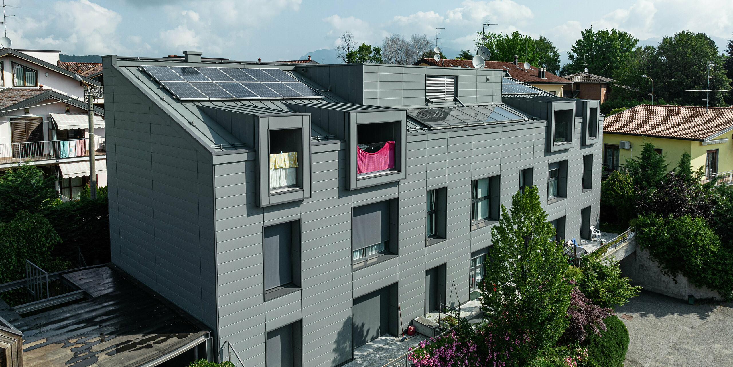 Vue de dessus du complexe résidentiel moderne de Faloppio, en Italie, avec une élégante façade en bardage PREFA et un toit équipé de modules photovoltaïques. Le Siding en aluminium durable, la production d'énergie respectueuse de l'environnement grâce à la puissance du soleil et l'apparence moderne créent un ensemble harmonieux. La combinaison d’une architecture esthétique et d’une technologie durable fait du bâtiment un excellent exemple de construction résidentielle contemporaine.