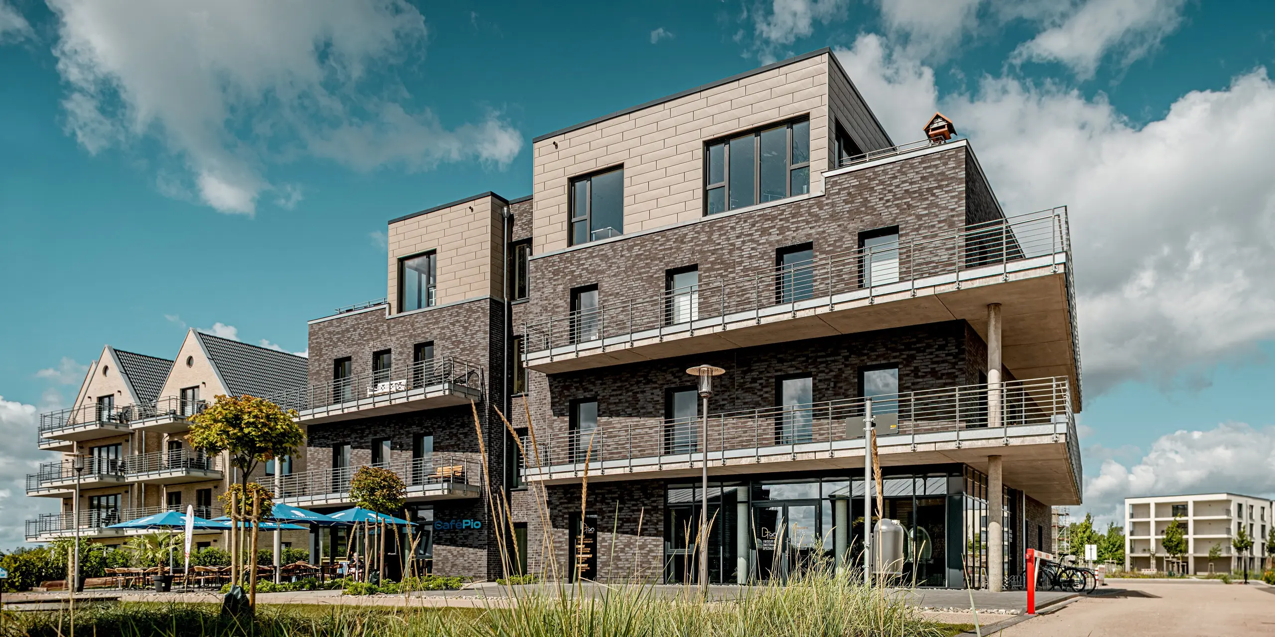 La photo présente un bâtiment du complexe résidentiel « Hafen zur Freiheit » (Port vers la liberté), caractérisé par une façade en briques sombres contrastant avec le dernier étage en retrait, revêtu de panneaux Siding.X PREFA couleur bronze élégant. Les surfaces en aluminium texturé confèrent à la partie supérieure une profondeur moderne et sophistiquée, créant un contraste saisissant avec la maçonnerie traditionnelle. De larges balcons, des lignes audacieuses et de nombreuses baies vitrées soulignent l'architecture ouverte. Résistants aux intempéries et durables, les panneaux PREFA offrent une protection pérenne et sont parfaitement adaptés à cet environnement côtier exposé.