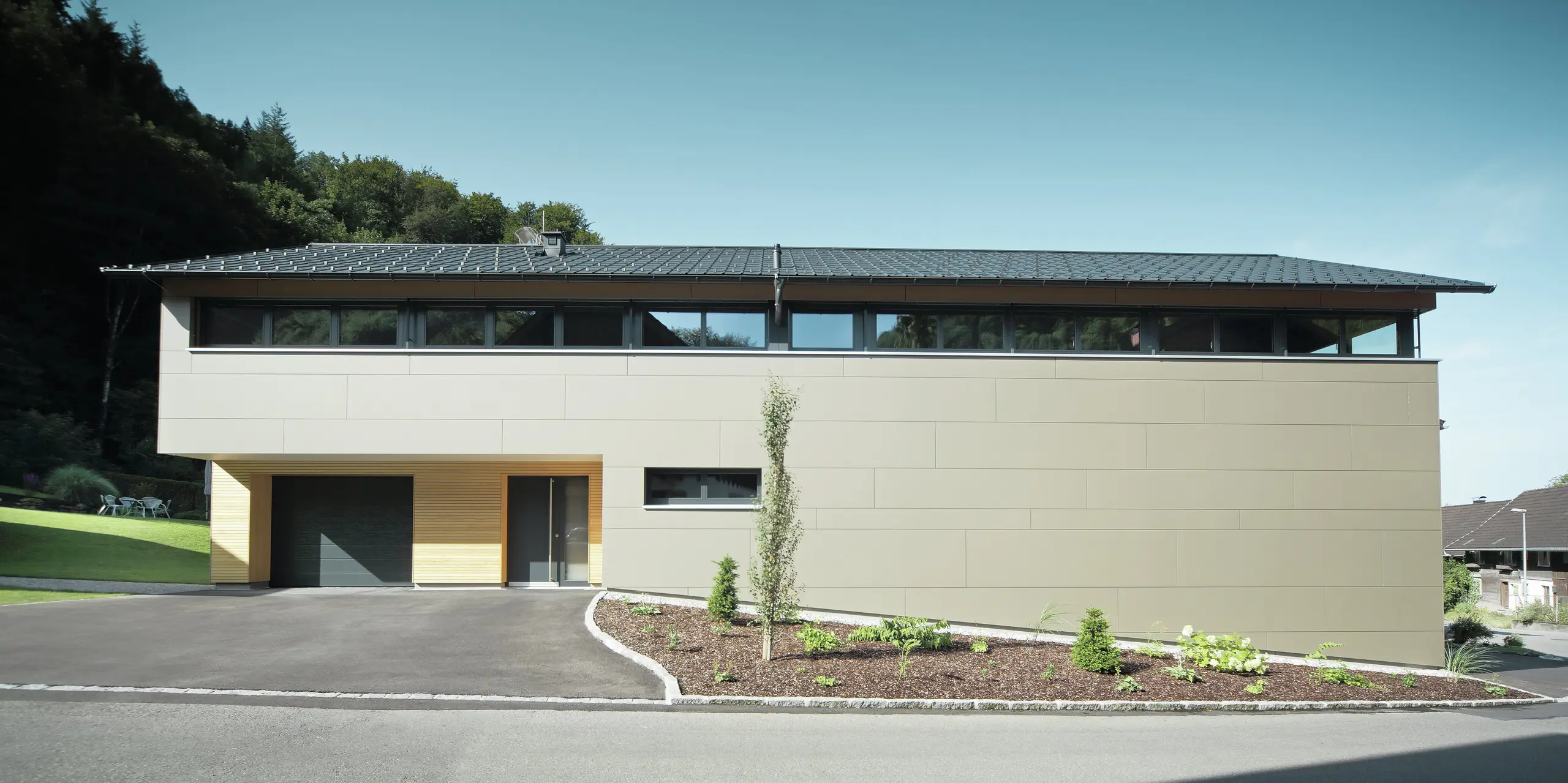 Das Einfamilienhaus in Hohenems präsentiert sich in einer modernen und stilvollen Fassade aus PREFA Aluminium Verbundplatten in Bronzemetallic, mit einem darüberliegenden Dach aus PREFA Dachplatten in P.10 Anthrazit. Die klare Linienführung und das durchdachte Farbkonzept des Designs integrieren das Gebäude nahtlos in die natürliche Umgebung. Die Gestaltung zeigt die Kombination aus Funktionalität und Ästhetik, die PREFA Produkte für zeitgemäße Wohnarchitektur bieten.