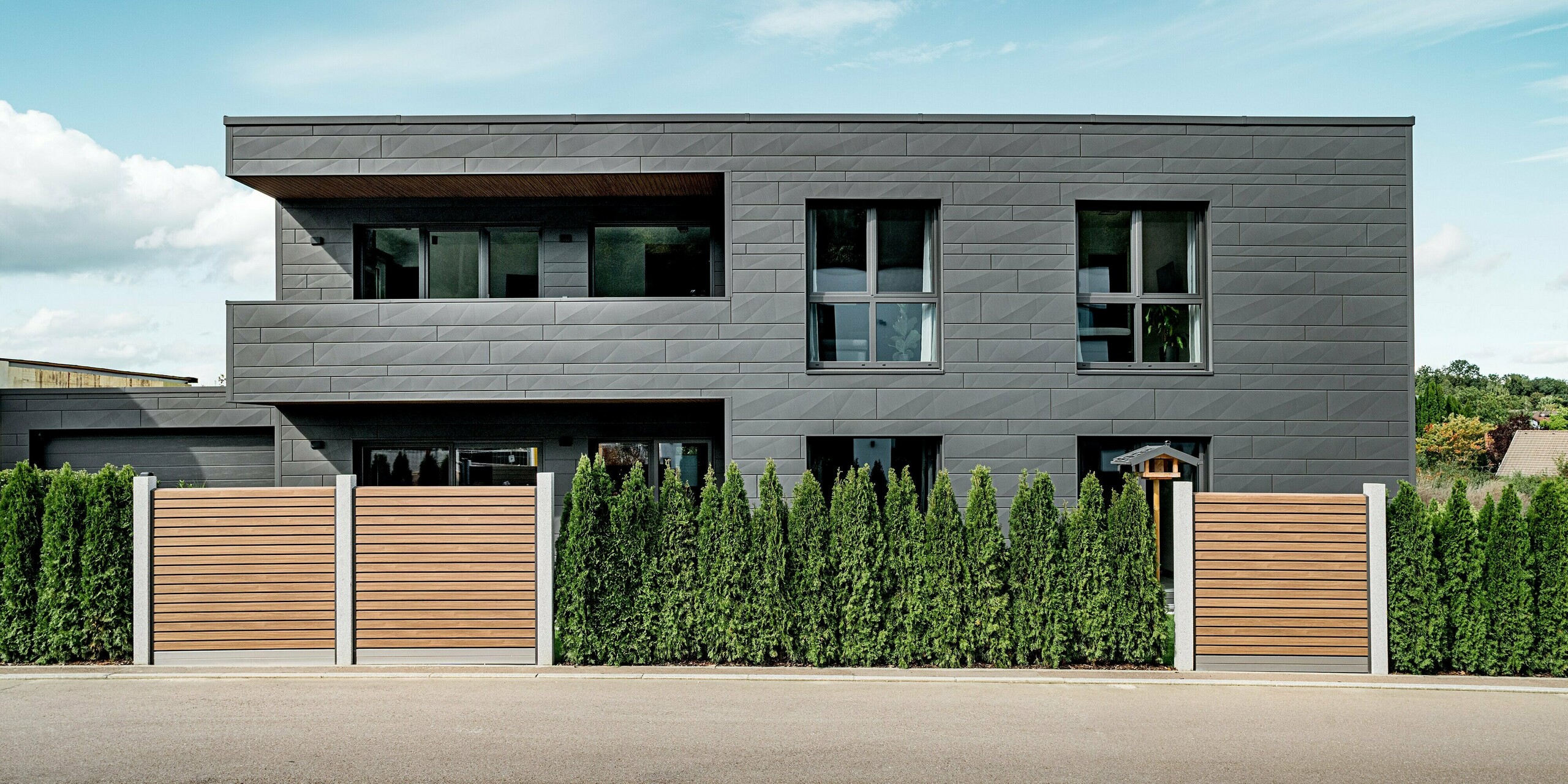 Straßenansicht eines modernen Einfamilienhauses in Heidenheim mit einer vollständig mit PREFA Siding.X Aluminium in der Farbe P.10 Dunkelgrau verkleideten Fassade. Die klar strukturierten Paneele mit unterschiedlichen Breiten und markanten Kantungen erzeugen eine dynamische, geometrische Fassadenwirkung. Im Vordergrund verläuft eine dichte Thujenhecke, die durch Holzzaunelemente mit vertikaler Lattung ergänzt wird. Diese bilden einen warmen Kontrast zur kühlen Metallarchitektur. Große Fensteröffnungen spiegeln Himmel und Umgebung. Die langlebige PREFA Aluminiumoberfläche sorgt für dauerhaften Schutz und eine moderne Optik.