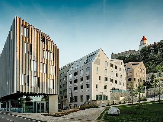 Dans le quartier de Vydrica à Bratislava, l'architecture moderne se mêle harmonieusement au contexte historique. Au premier plan, un bâtiment sculptural à la façade verticale captive le regard, tandis qu'à l'arrière-plan, des toitures à forte pente, revêtues d'ardoises en aluminium PREFA blanc et brun sable, créent des accents précis. La structure claire des toitures, intégrant des fenêtres, contraste fortement avec le château qui se dresse à flanc de colline. L'aluminium PREFA impressionne par sa grande malléabilité, sa durabilité et son extrême résistance aux intempéries – un matériau idéal pour les projets urbains exigeants sur le plan architectural.
