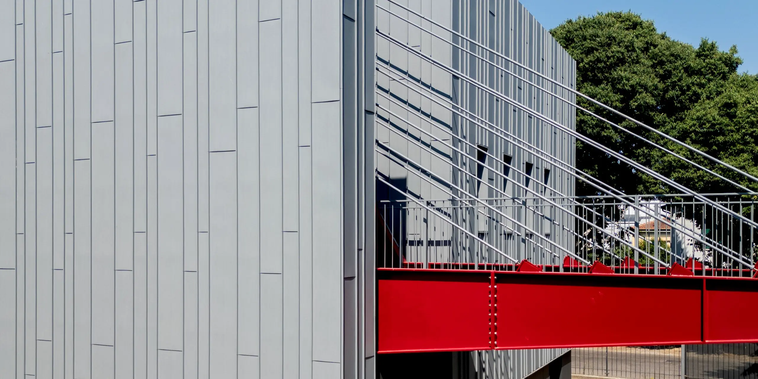 Une vue saisissante de la Scuola Arte e Mestieri F. Bertazzoni à Suzzara révèle un spectaculaire pont rouge vif enjambant deux ailes du bâtiment, ainsi que la façade verticale en aluminium PREFA, d'un gris quartz, témoignant d'une grande précision. La pose impeccable des panneaux PREFALZ, avec leurs joints debout caractéristiques, souligne avec élégance la rigueur architecturale de l'édifice. Les câbles tendus du pont projettent des ombres fascinantes sur la façade. Ici, les produits en aluminium PREFA démontrent leur résistance durable aux intempéries, leur stabilité dimensionnelle et leur esthétique intemporelle.