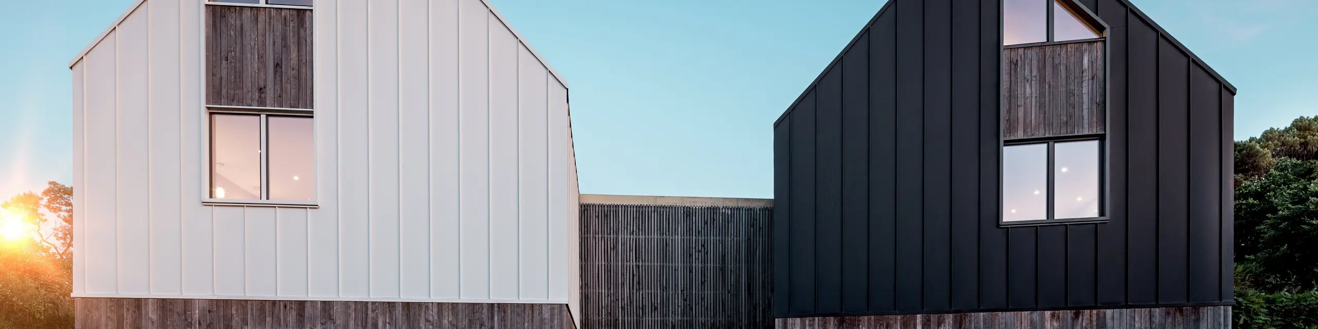 La façade du complexe résidentiel Le Rhune présente deux bâtiments décalés, à l'architecture identique, qui se distinguent par leurs contrastes de blanc et de noir. La pureté des lignes des toitures à deux pans et le revêtement continu en aluminium PREFALZ confèrent aux bâtiments une allure sculpturale et mettent en valeur la précision des joints debout. De chaleureux jeux de lumière, filtrés par les larges fenêtres, créent un contraste saisissant avec l'architecture minimaliste. L'aluminium PREFALZ, résistant aux intempéries et durable, assure la cohésion de la toiture et de la façade, formant une enveloppe de bâtiment moderne et harmonieuse, d'une grande qualité esthétique.