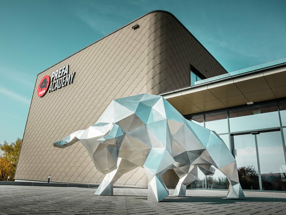 Une imposante sculpture de taureau en aluminium brillant de 2 mm caractérise la zone d'entrée saisissante du nouveau site PREFA à Törökbálint. La façade moderne avec des losanges de façade PREFA 44 × 44 en bronze confère au bâtiment nouvellement construit une enveloppe extérieure élégante, résistante aux intempéries et durable. Les portes coulissantes en verre créent une atmosphère lumineuse dès l'entrée dans le bâtiment. Le logo PREFA Academy, clairement visible sur la façade, souligne le caractère fort du bâtiment de l'entreprise.