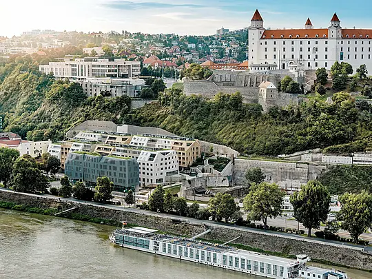 Vue panoramique sur le quartier de Vydrica à Bratislava, surplombant le château et le Danube : l’architecture aux multiples facettes séduit par ses toitures végétalisées, ses toits à forte pente et ses puits de lumière parfaitement intégrés. Les tuiles de toiture en aluminium PREFA, disponibles en blanc, brun sable et bronze, unifient les bâtiments en un ensemble harmonieux et s’intègrent parfaitement au paysage historique. Durables, légères et extrêmement résistantes aux intempéries, les solutions PREFA permettent des géométries de toiture complexes et garantissent une enveloppe de bâtiment durable, nécessitant peu d’entretien et d’une grande qualité esthétique.