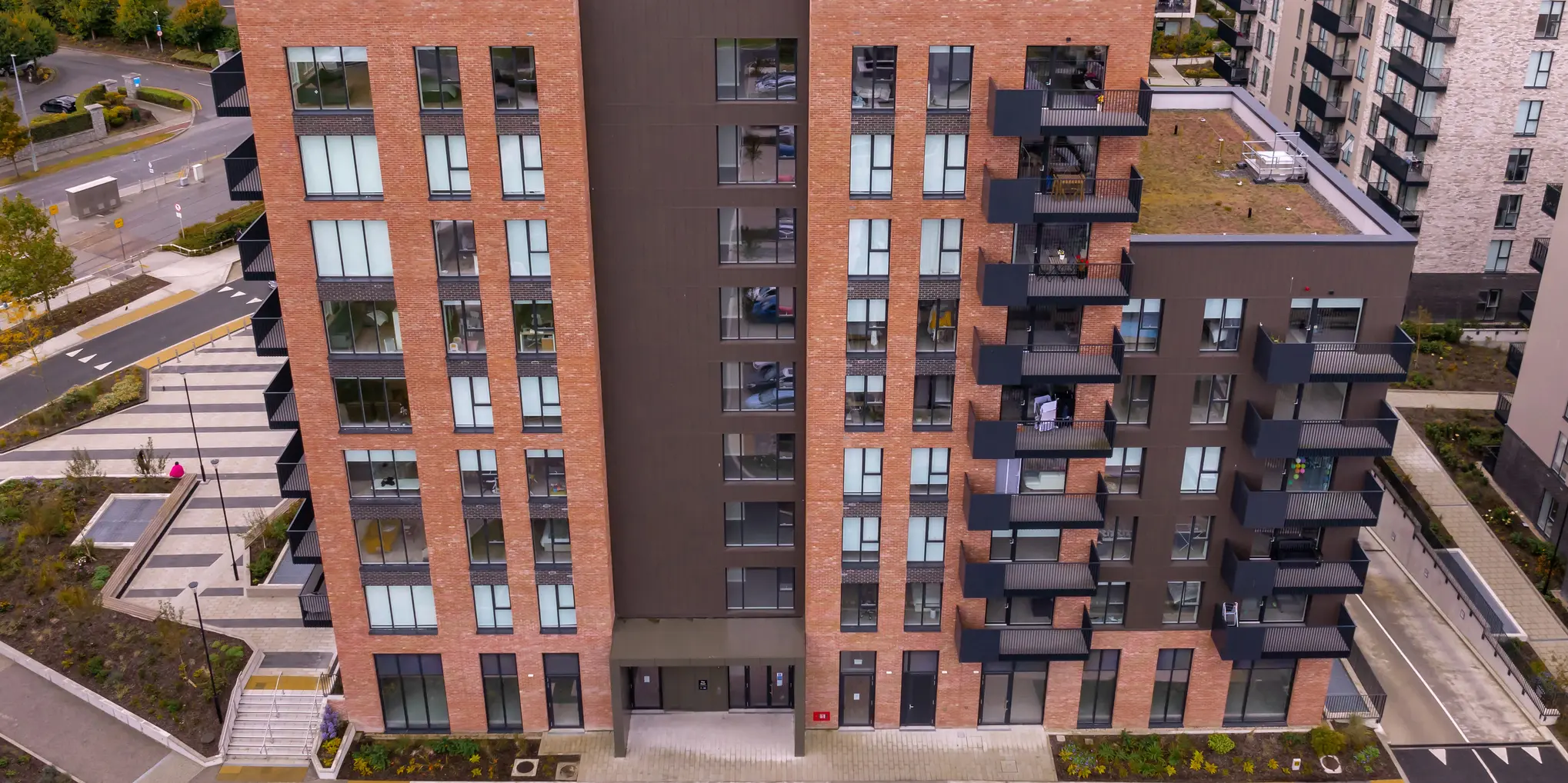 Vue plongeante sur les appartements Citywest à Dublin. La combinaison de façade en briques traditionnelles et de 700 m² de revêtements PREFA en P.10 brun s'intègre harmonieusement dans l'architecture moderne et l'environnement. Vous pouvez voir la zone d'entrée du complexe résidentiel avec des espaces verts bien entretenus autour du bâtiment.