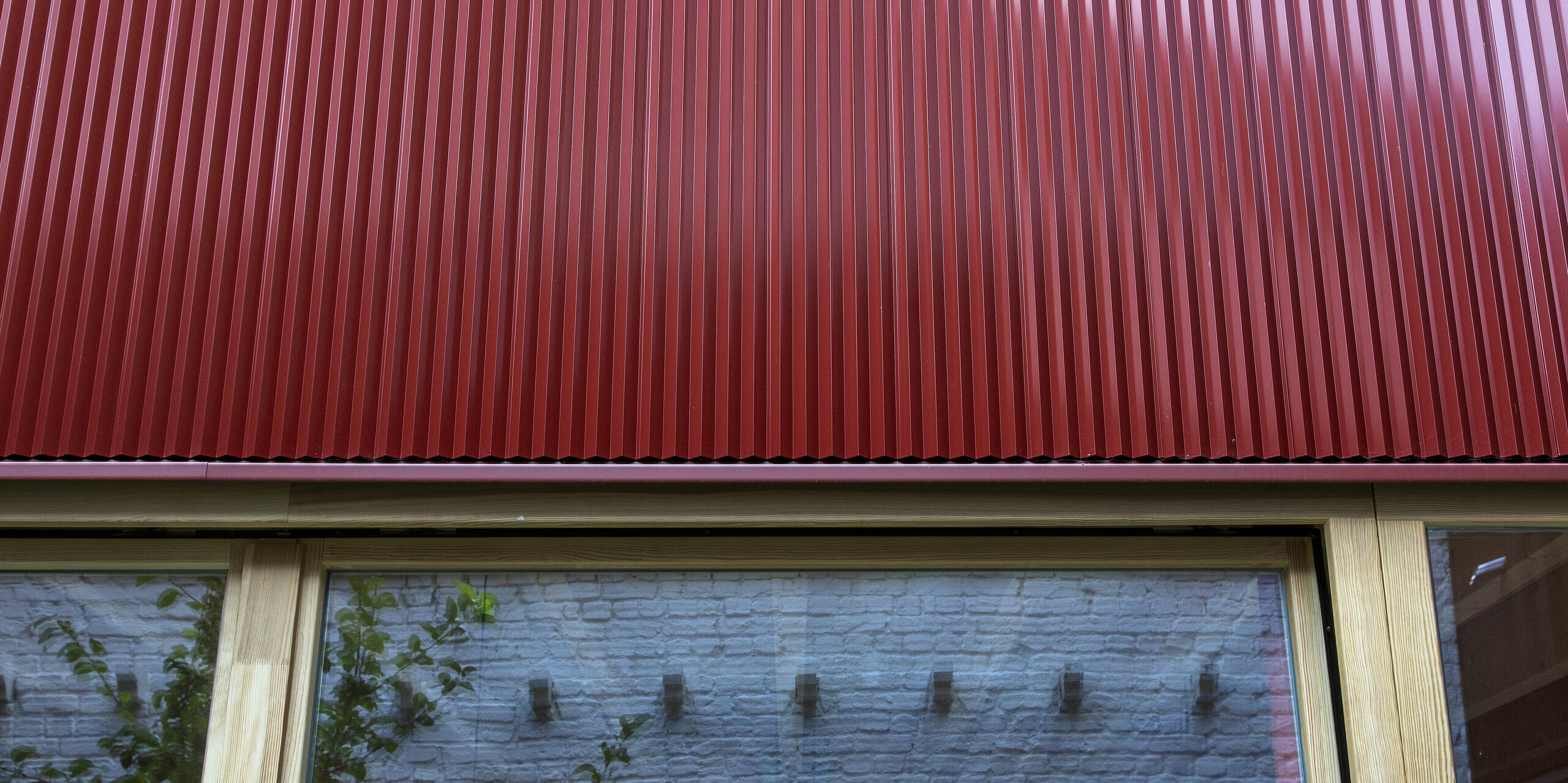 Gros plan sur le profil triangle PREFA rouge oxyde au-dessus d'une grande fenêtre en bois : la précision de la fabrication et la structure verticale de l'aluminium extrudé créent une façade moderne et remarquable. Ce profilé en tôle robuste résiste aux intempéries, est indéformable et apporte au bâtiment une touche architecturale de grande qualité.