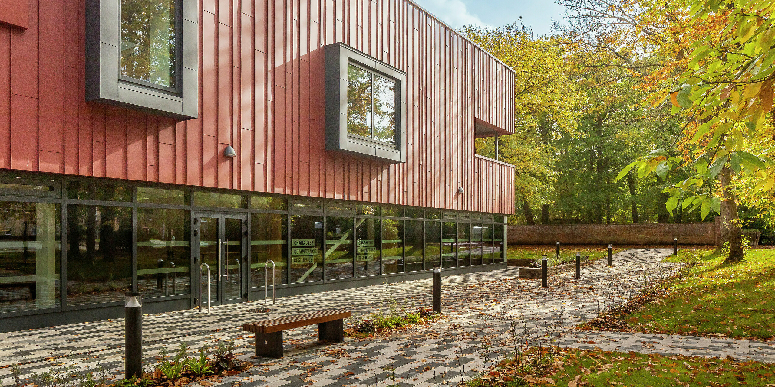 Une architecture harmonieuse dans un cadre verdoyant : La façade moderne à joint debout du Soldiers' Centre de Pirbright est réalisée en PREFALZ en P.10 rouge oxyde. Le revêtement robuste en aluminium s'intègre parfaitement dans l'environnement automnal. Les cadres de fenêtre en porte-à-faux remarquables en P.10 anthracite apportent des accents élégants. De grandes surfaces vitrées au rez-de-chaussée assurent transparence et ouverture. Le parvis pavé avec banc vous invite à vous attarder - une combinaison réussie de design, de fonctionnalité et de nature.