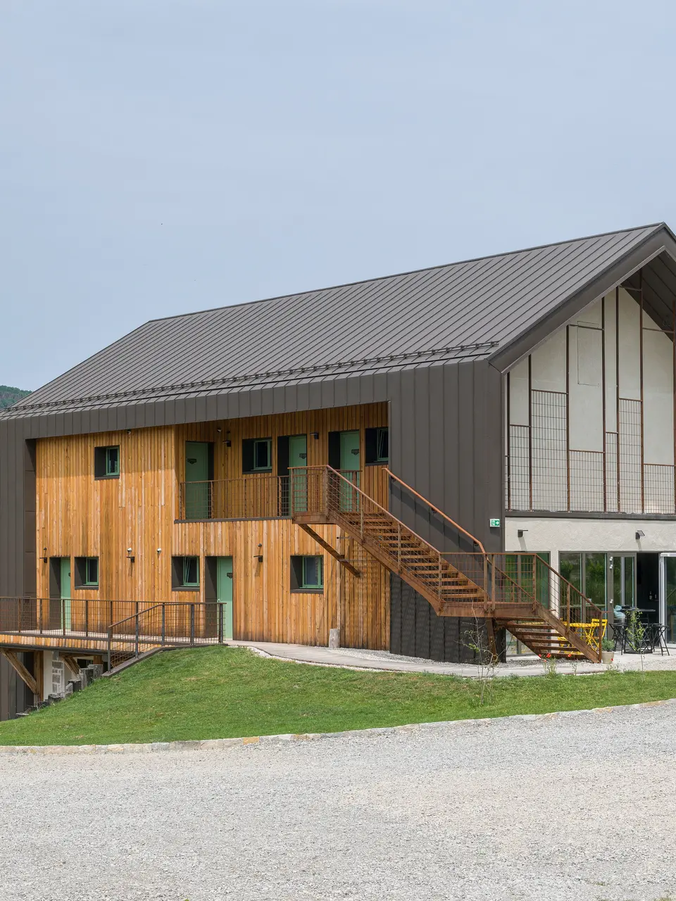 Vue d'angle d'un hôtel contemporain à deux étages avec une coque en aluminium PREFA et une façade en bois naturel. Le toit et certaines parties de la façade ont été revêtus de PREFALZ en brun P.10. Les caractéristiques remarquables du bâtiment sont de grandes fenêtres et un escalier extérieur qui mène à l'étage supérieur. L'Hôtel en Tilleul dispose également d'une terrasse au rez-de-chaussée. Le bâtiment se trouve dans un cadre rural idyllique surplombant des collines. La route d'accès en gravier est visible au bas de la photo.