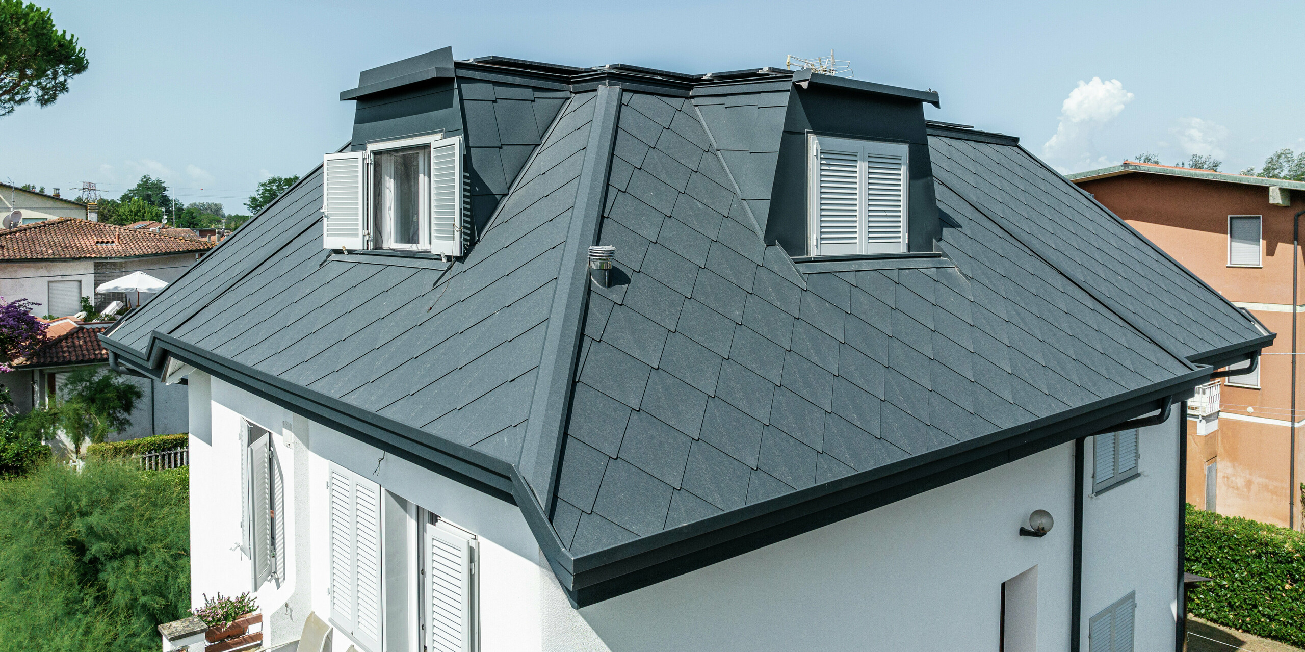 La finition précise du toit en losanges PREFA 44 × 44 en P.10 gris pierre sur la maison familiale de Montignoso est clairement visible dans ce gros plan. Le revêtement en aluminium de haute qualité assure un look élégant et moderne tout en offrant une résistance maximale aux conditions météorologiques extrêmes. Les élégantes lucarnes s'intègrent harmonieusement dans le toit et créent un espace de vie supplémentaire sous la couverture de toit robuste et durable. Une interaction parfaite entre design et fonctionnalité.