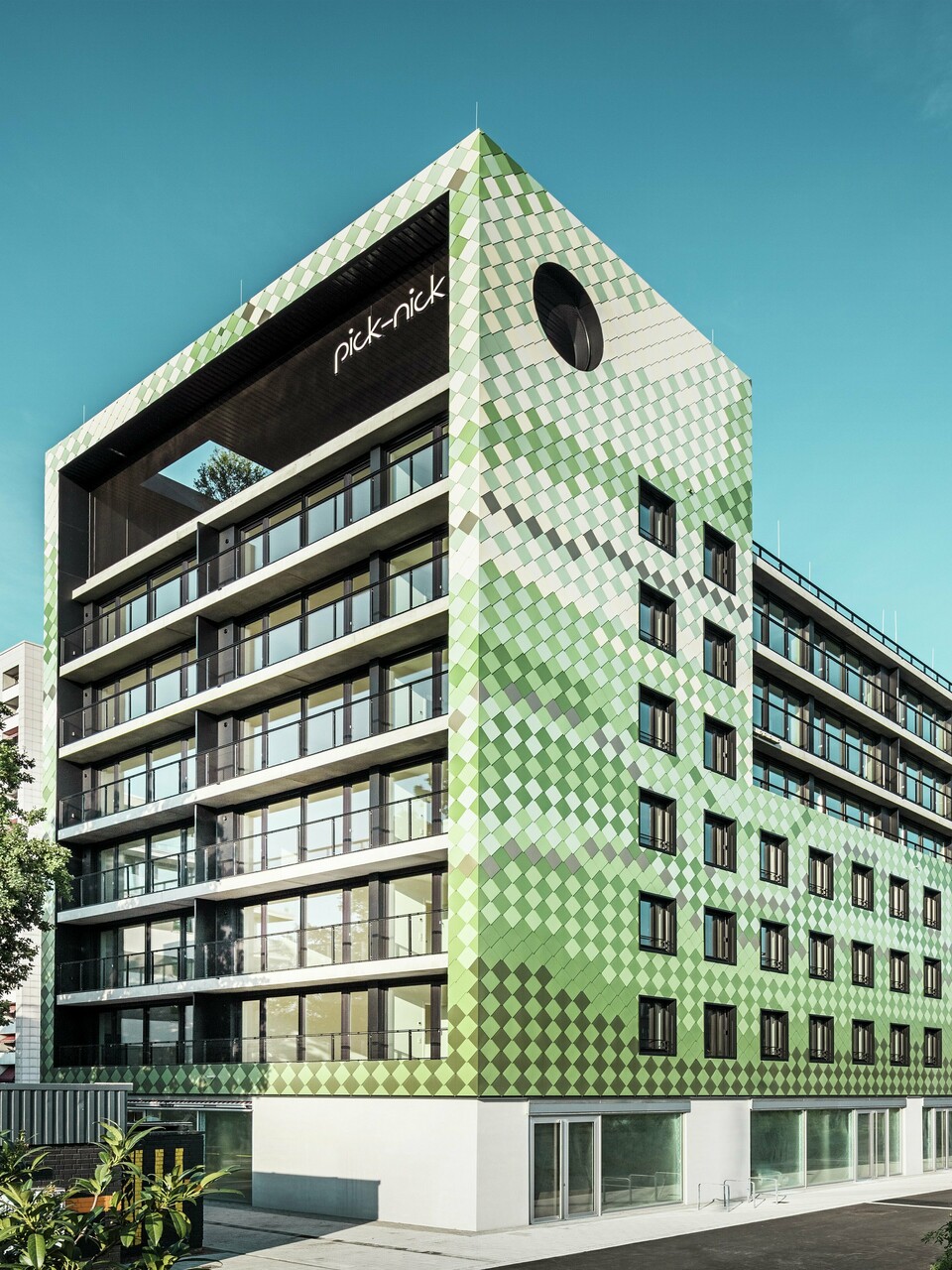 Bâtiment résidentiel et commercial « Pick-Nick » à Dresde avec une façade moderne en panneaux d'aluminium PREFA dans différents tons verts et beiges. La façade distinctive du bâtiment combine des lignes claires avec un jeu de couleurs dynamique et est en outre enrichie de grandes surfaces vitrées. Les accents architecturaux comprennent l'inscription « Pick-Nick » sur le bord supérieur du bâtiment et le jardin sur le toit vert.
