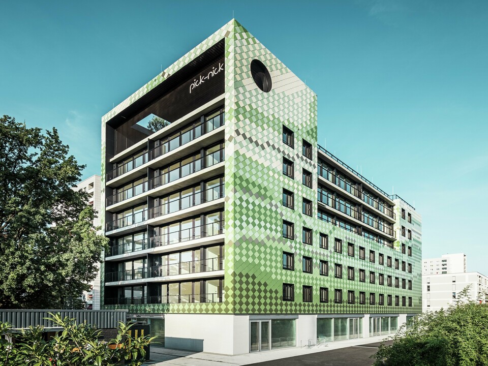 Bâtiment résidentiel et commercial « Pick-Nick » à Dresde avec une façade moderne en panneaux d'aluminium PREFA dans différents tons verts et beiges. La façade distinctive du bâtiment combine des lignes claires avec un jeu de couleurs dynamique et est en outre enrichie de grandes surfaces vitrées. Les accents architecturaux comprennent l'inscription « Pick-Nick » sur le bord supérieur du bâtiment et le jardin sur le toit vert.