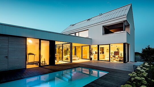 L'image présente une maison individuelle moderne à Freistadt, dotée d'une façade PREFALZ d'un blanc immaculé. La façade en aluminium, aux plis précis, confère au bâtiment une élégance géométrique épurée, en harmonie avec les généreuses baies vitrées et le toit plat. L'utilisation du matériau PREFALZ garantit non seulement une esthétique intemporelle, mais aussi une grande résistance aux intempéries et une durabilité exceptionnelle. L'intérieur décloisonné, avec sa vue sur la piscine, est parfaitement mis en valeur par les lignes épurées et le design minimaliste, faisant de cette maison une solution d'habitation contemporaine et durable.