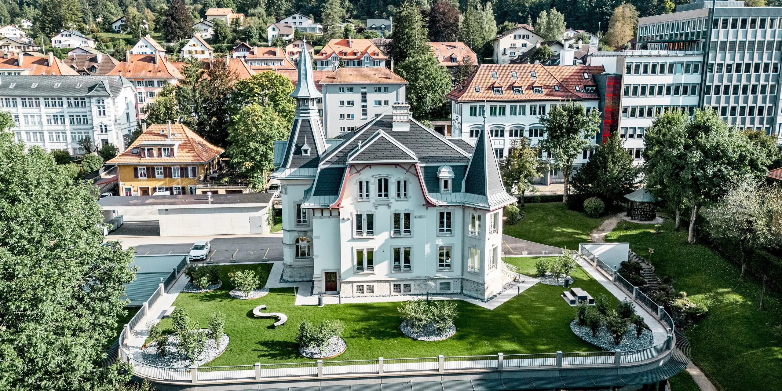 Weitläufige Gesamtansicht der Manufacture Horlogère du Locle in der Schweiz, eingebettet in eine grüne Parklandschaft und umgeben von historischer Bebauung. Im Fokus steht die ikonische Dacharchitektur mit eleganten Türmen, steilen Flächen und geschwungenen Formen. PREFA Dachschindeln in P.10 Schwarz betonen präzise die Geometrie, während PREFALZ in Patinagrau fließende Übergänge ermöglicht. Das leichte Aluminiumdach überzeugt durch Langlebigkeit, Witterungsbeständigkeit und maximale Gestaltungsfreiheit bei komplexen Dachformen.