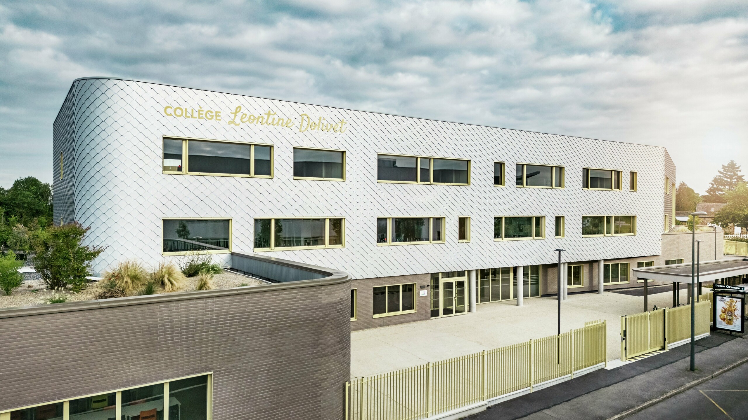 Vue de face du Collège Léontine Dolivet à Cesson-Sévigné, dont la façade moderne en aluminium est composée de losanges PREFA argent métallisé. La surface irisée confère au bâtiment une impression de légèreté, tandis que les lignes épurées et les larges fenêtres assurent une transparence optimale. Des encadrements de fenêtres jaunes apportent des touches de dynamisme. Au premier plan, on aperçoit une place lumineuse, une clôture et la rue adjacente. La solution de façade PREFA, durable et résistante, allie esthétique, résistance aux intempéries et durabilité – un véritable atout architectural, témoignant d'une élégance raffinée.