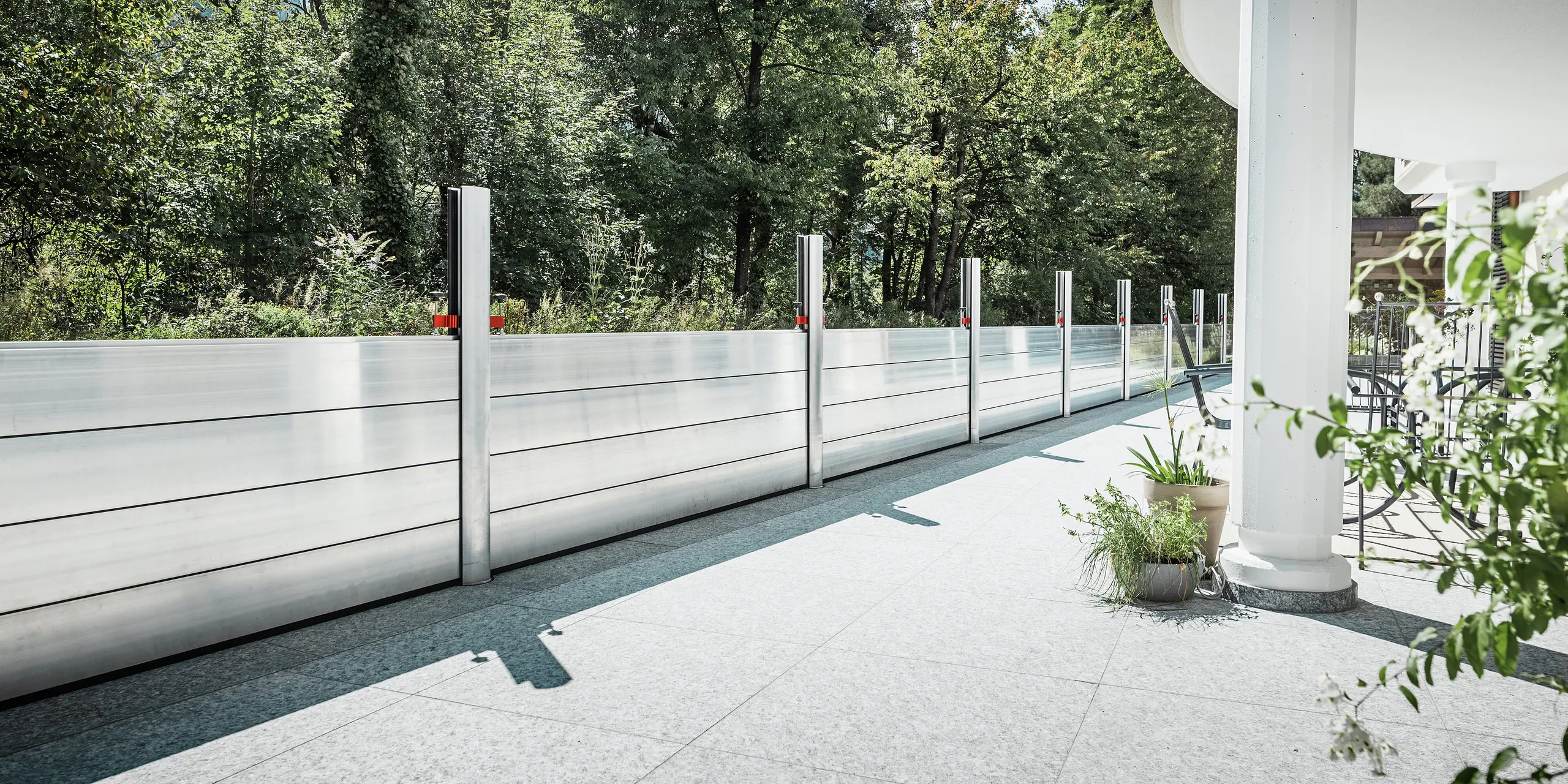 Vue depuis une élégante terrasse abritée, équipée d'un système de protection contre les crues PREFA. Les batardeaux en aluminium s'étendent en ligne droite le long de la limite de propriété jusqu'à la forêt adjacente, offrant une protection efficace contre les inondations. Les éléments en aluminium brillant s'intègrent harmonieusement au design moderne de la maison. Au premier plan : colonnes blanches, jardinières et mobilier de jardin, soulignant l'atmosphère chaleureuse.