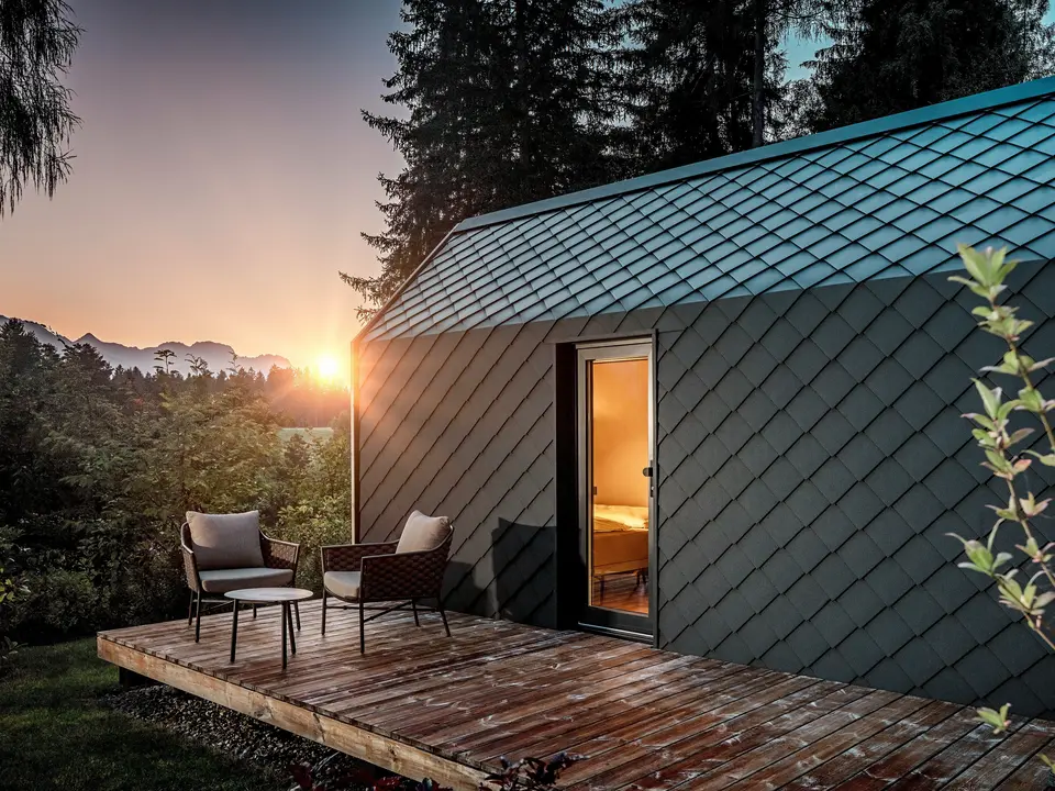 Au coucher du soleil, une minuscule maison minimaliste nichée dans le Secret Koi Garden, sur les rives du lac Natterer, se dévoile. Sa terrasse en bois offre une vue imprenable sur la forêt. Toit et façade sont revêtus de petits losanges en aluminium PREFA P.10 anthracite, dont la surface texturée reflète avec élégance la douce lumière. Durable et résistant aux intempéries, l'aluminium PREFA protège efficacement la structure et crée un contraste saisissant avec le bois, la nature environnante et la lumière tamisée.