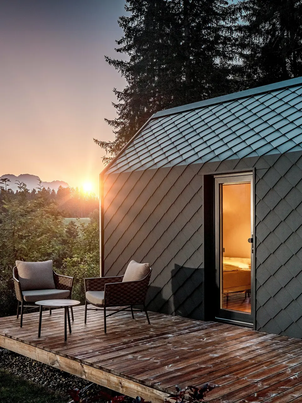 Au coucher du soleil, une minuscule maison minimaliste nichée dans le Secret Koi Garden, sur les rives du lac Natterer, se dévoile. Sa terrasse en bois offre une vue imprenable sur la forêt. Toit et façade sont revêtus de petits losanges en aluminium PREFA P.10 anthracite, dont la surface texturée reflète avec élégance la douce lumière. Durable et résistant aux intempéries, l'aluminium PREFA protège efficacement la structure et crée un contraste saisissant avec le bois, la nature environnante et la lumière tamisée.
