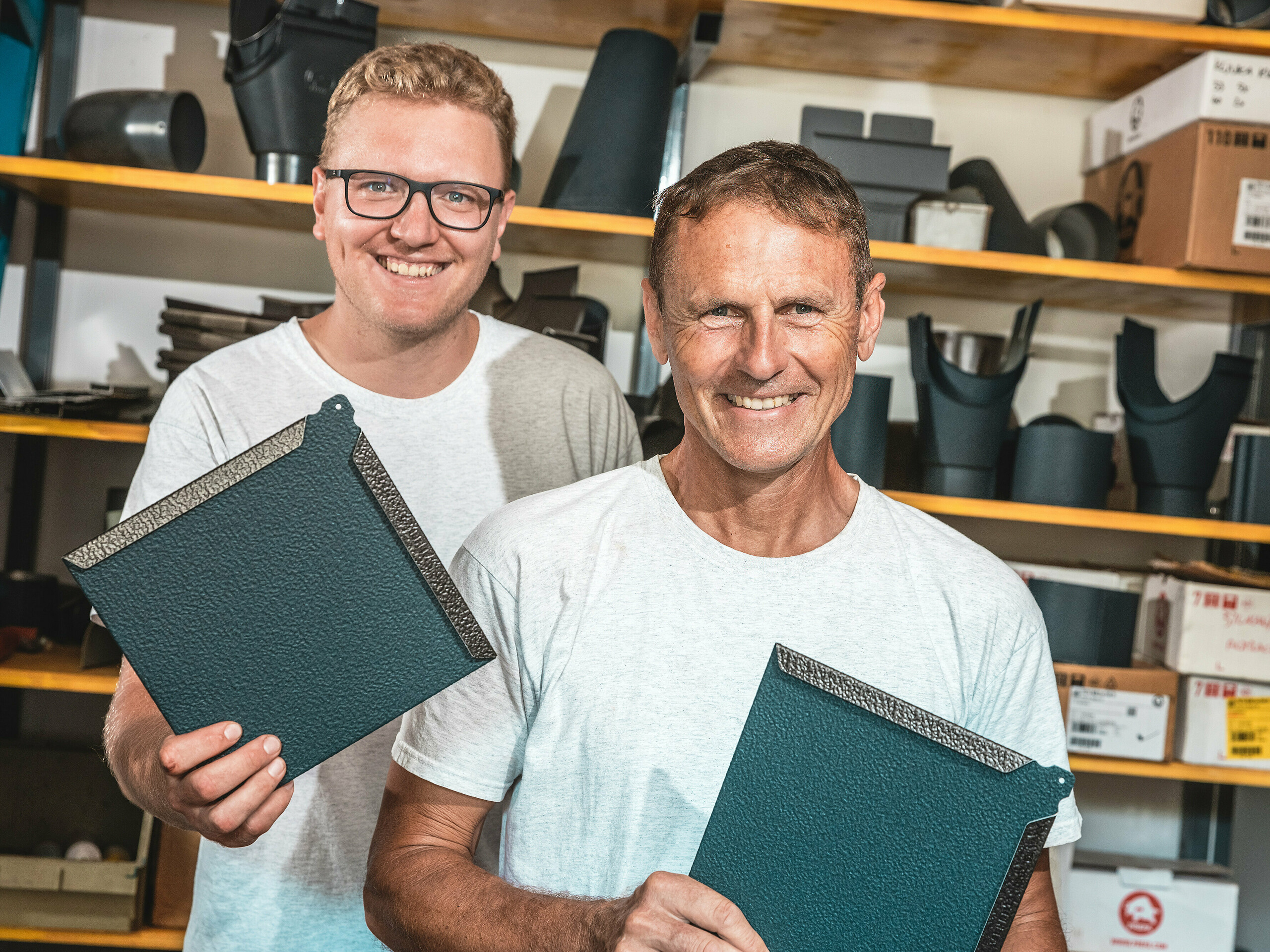 Dans un atelier, deux artisans présentent des losanges en aluminium PREFA, coloris P.10 anthracite. Au premier plan, Janez Kralj, maître ferblantier slovène, se tient derrière lui, un jeune membre de son équipe. À l'arrière-plan, des étagères présentent des composants et accessoires pour systèmes de toiture et de façade. La surface finement texturée des losanges en aluminium PREFA témoigne de leur grande qualité, de leur durabilité et de leur fabrication soignée. Ils sont parfaitement adaptés aux projets architecturaux exigeants, tels que les mini-maisons, où résistance, légèreté et design moderne sont essentiels.