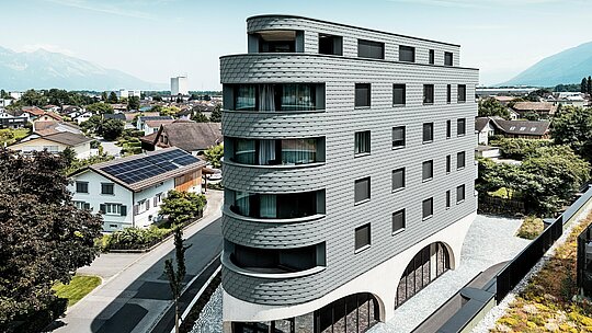 Vue aérienne de la Bernerhaus à Buchs, reconnaissable à sa forme triangulaire et ses balcons aux courbes douces. La façade en bardeau gris souris PREFA P.10 enveloppe le bâtiment comme une peau d'aluminium aux écailles, créant un jeu d'ombre et de lumière saisissant. Au-dessus du socle en béton massif aux élégantes arcades, la façade en aluminium PREFA, légère et résistante aux intempéries, se distingue par sa précision, sa durabilité et son faible besoin d'entretien – un élément architectural moderne qui attire le regard dans le paysage alpin.