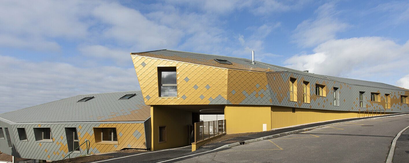 Vue de la route de l'école primaire. La façade a été réalisée en losange 29x29 PREFA dans les teintes Gris souris et Mayagold.