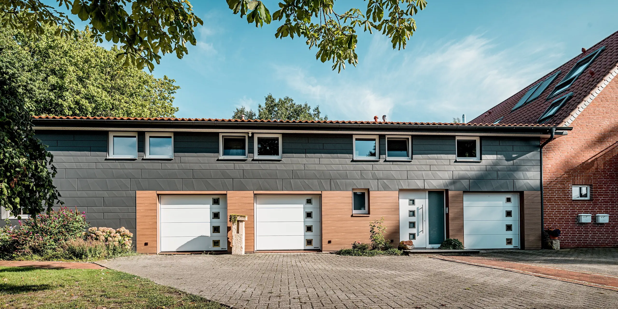 Vue d'ensemble d'une maison individuelle rénovée, dotée d'une façade moderne en aluminium PREFA aux lignes horizontales épurées. La façade associe le revêtement PREFA Siding.X P.10 gris sombre dans sa partie supérieure au revêtement PREFA Siding.X noyer foncé à sa base et à l'entrée, créant un contraste saisissant entre élégance et chaleur du bois. Les bords distinctifs des panneaux Siding.X créent de magnifiques jeux d'ombre et de lumière, tandis que la finition bois apporte des accents ciblés. Les portes de garage blanches et les fenêtres aux dimensions précises soulignent l'aspect moderne. Durables, résistants aux intempéries et nécessitant peu d'entretien, les panneaux en aluminium PREFA font de cette rénovation de façade une solution durable, de haute qualité et architecturalement expressive.