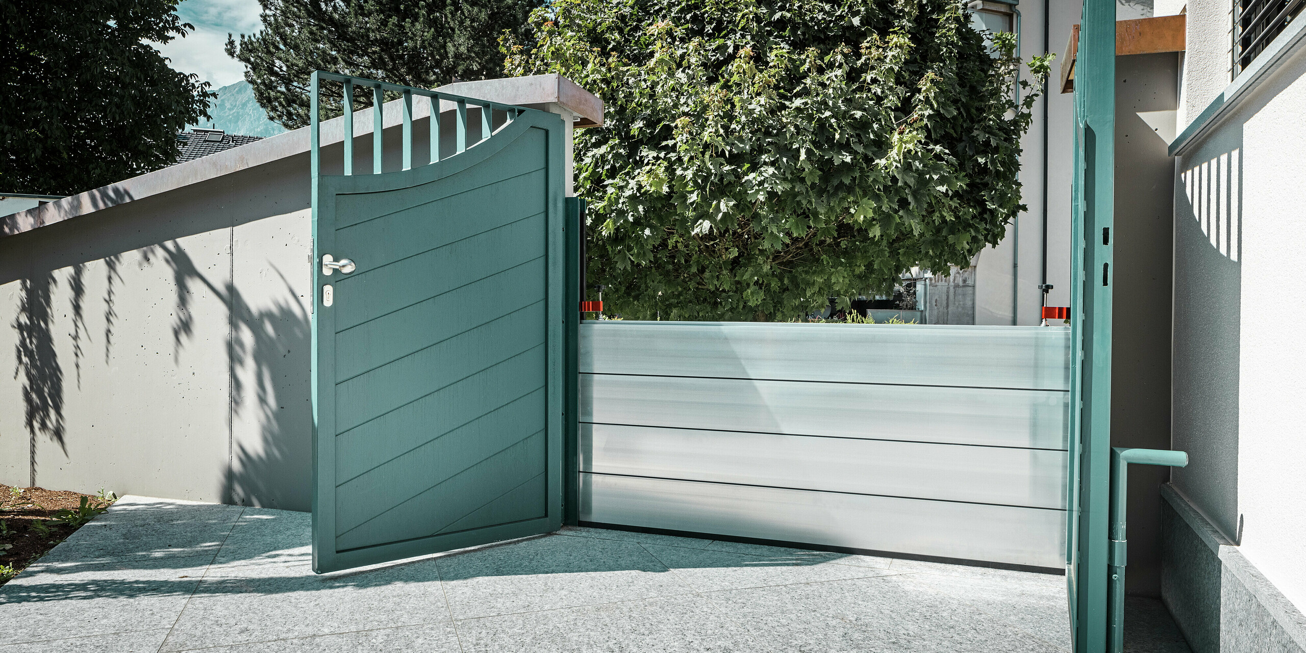 Portail de jardin ouvert avec système de protection contre les crues PREFA intégré devant une maison individuelle à Schwaz, au Tyrol. Les batardeaux en aluminium sécurisent l'entrée contre le mur de la maison et assurent une protection efficace du bien en cas d'inondation. Le système s'intègre discrètement à l'architecture existante et souligne l'alliance parfaite entre sécurité fonctionnelle et esthétique.