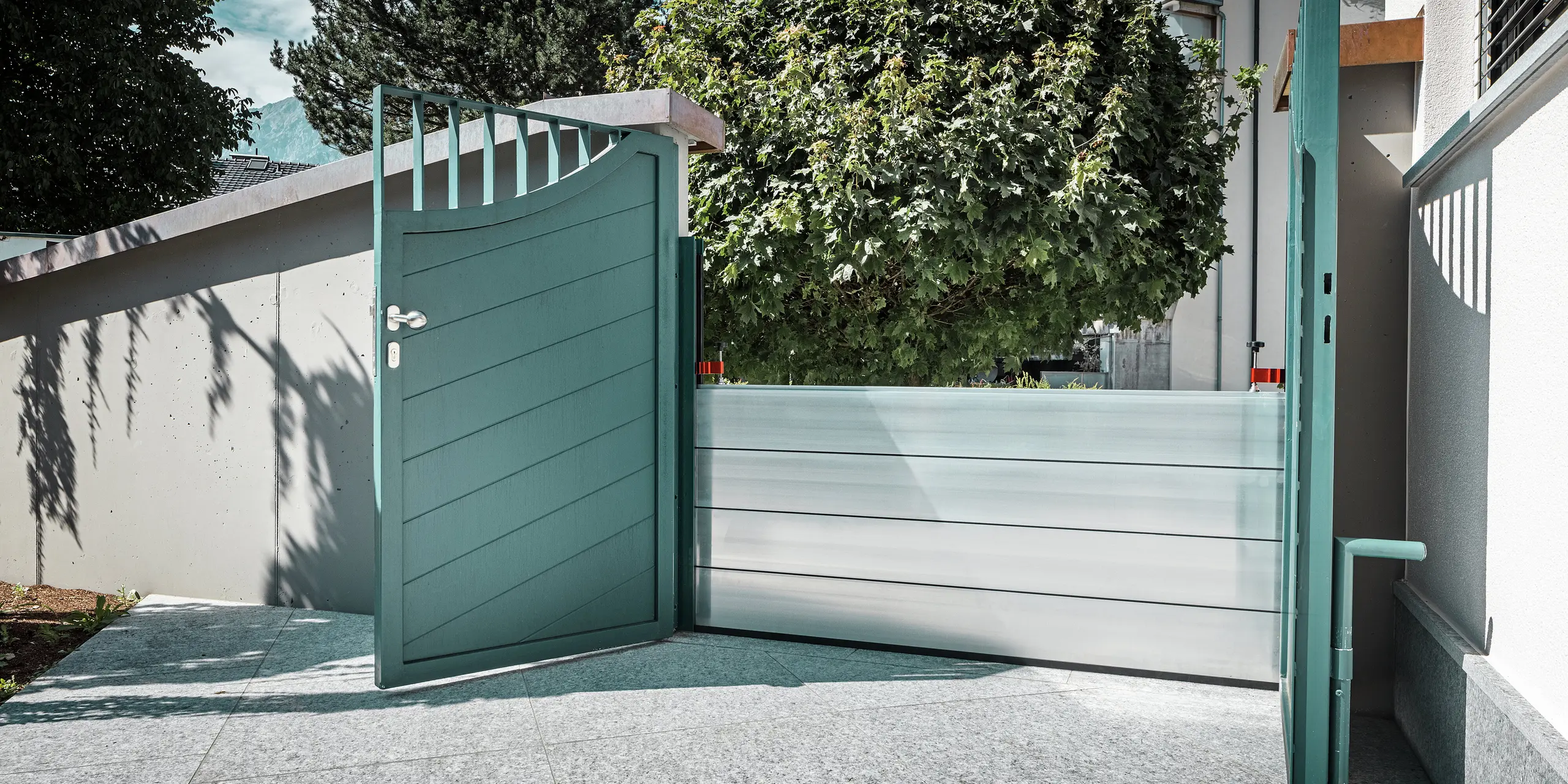 Portail de jardin ouvert avec système de protection contre les crues PREFA intégré devant une maison individuelle à Schwaz, au Tyrol. Les batardeaux en aluminium sécurisent l'entrée contre le mur de la maison et assurent une protection efficace du bien en cas d'inondation. Le système s'intègre discrètement à l'architecture existante et souligne l'alliance parfaite entre sécurité fonctionnelle et esthétique.