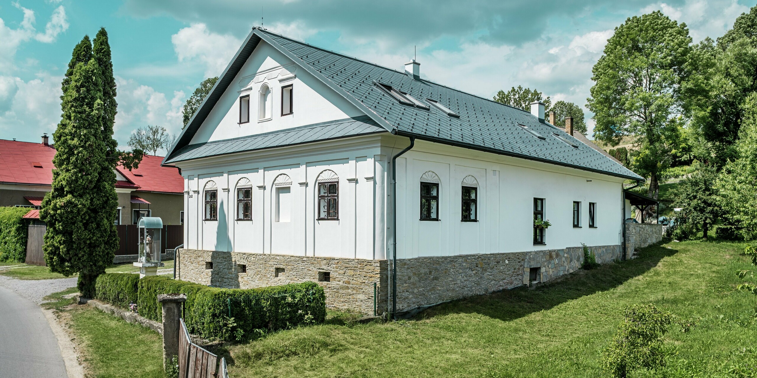 La photo prise depuis la rue montre la façade et le côté d'une maison d'artisan rénovée à Klin, en Slovaquie. Elle présente une toiture moderne en losanges PREFA 44 x 44, coloris Gris Pierre P.10. La vue latérale révèle l'élégant toit à pignon et les puits de lumière parfaitement intégrés, créant une atmosphère lumineuse et aérée. Les losanges en aluminium s'harmonisent avec la façade blanche traditionnelle et la maçonnerie en pierre naturelle, conférant à la maison un caractère à la fois contemporain et classique. Grâce à ses atouts tels que sa durabilité, sa légèreté et sa résistance à la corrosion, cette solution de toiture est non seulement esthétique, mais aussi très fonctionnelle et durable.