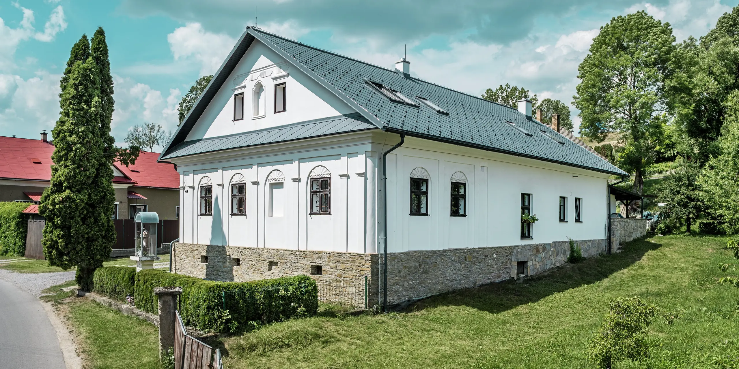 La photo prise depuis la rue montre la façade et le côté d'une maison d'artisan rénovée à Klin, en Slovaquie. Elle présente une toiture moderne en losanges PREFA 44 x 44, coloris Gris Pierre P.10. La vue latérale révèle l'élégant toit à pignon et les puits de lumière parfaitement intégrés, créant une atmosphère lumineuse et aérée. Les losanges en aluminium s'harmonisent avec la façade blanche traditionnelle et la maçonnerie en pierre naturelle, conférant à la maison un caractère à la fois contemporain et classique. Grâce à ses atouts tels que sa durabilité, sa légèreté et sa résistance à la corrosion, cette solution de toiture est non seulement esthétique, mais aussi très fonctionnelle et durable.
