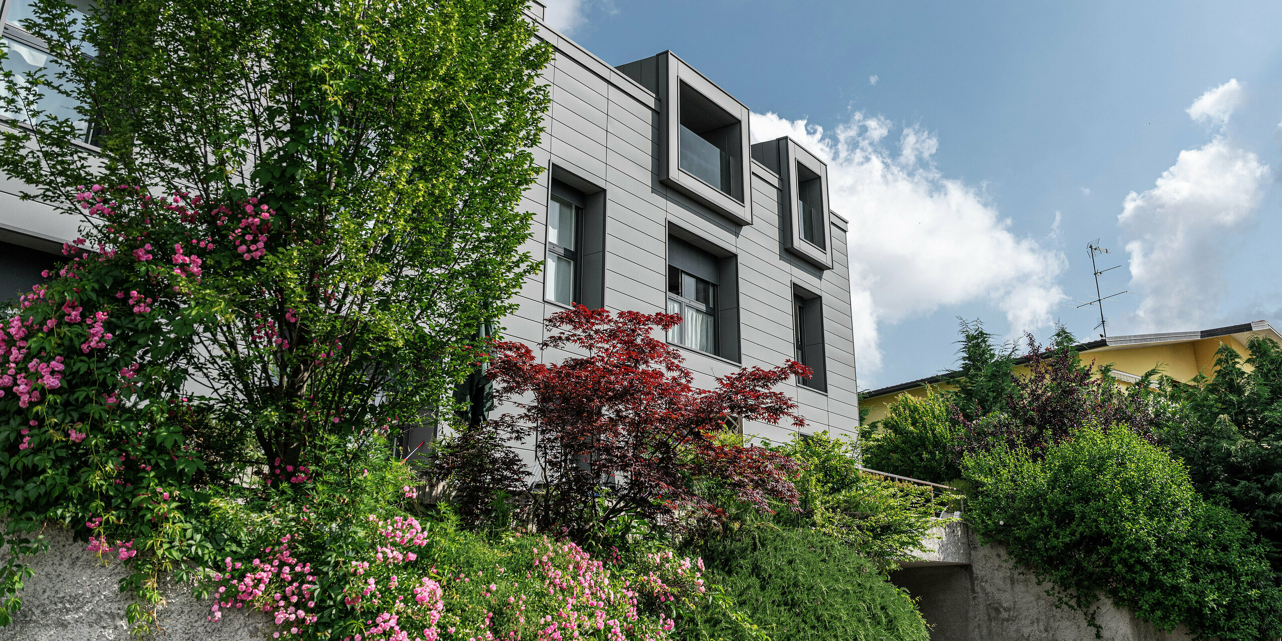 Complexe résidentiel moderne à Faloppio, en Italie, avec une façade élégante en Sidings PREFA. Les fenêtres en porte-à-faux offrent un attrait architectural unique, tandis que les plantes et les fleurs environnantes rehaussent la beauté naturelle de la propriété. Le revêtement en aluminium durable de PREFA offre une solution esthétique et fonctionnelle dans toutes les conditions météorologiques.