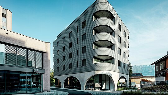 Cette maison bernoise moderne, située à Buchs, séduit par sa forme triangulaire arrondie et sa façade continue en bardage PREFA P.10 Gris souris. La surface en aluminium, à l'aspect d'écailles, souligne les lignes douces et les courbes profondes du balcon, tandis que le socle en béton massif, orné d'élégantes arcades, crée un contraste saisissant. Se détachant sur un fond alpin, au cœur d'une petite rue commerçante, la façade en aluminium PREFA, légère, résistante aux intempéries et durable, devient un véritable atout architectural : fabriquée avec précision, elle nécessite peu d'entretien et s'adapte parfaitement aux courbes les plus exigeantes.