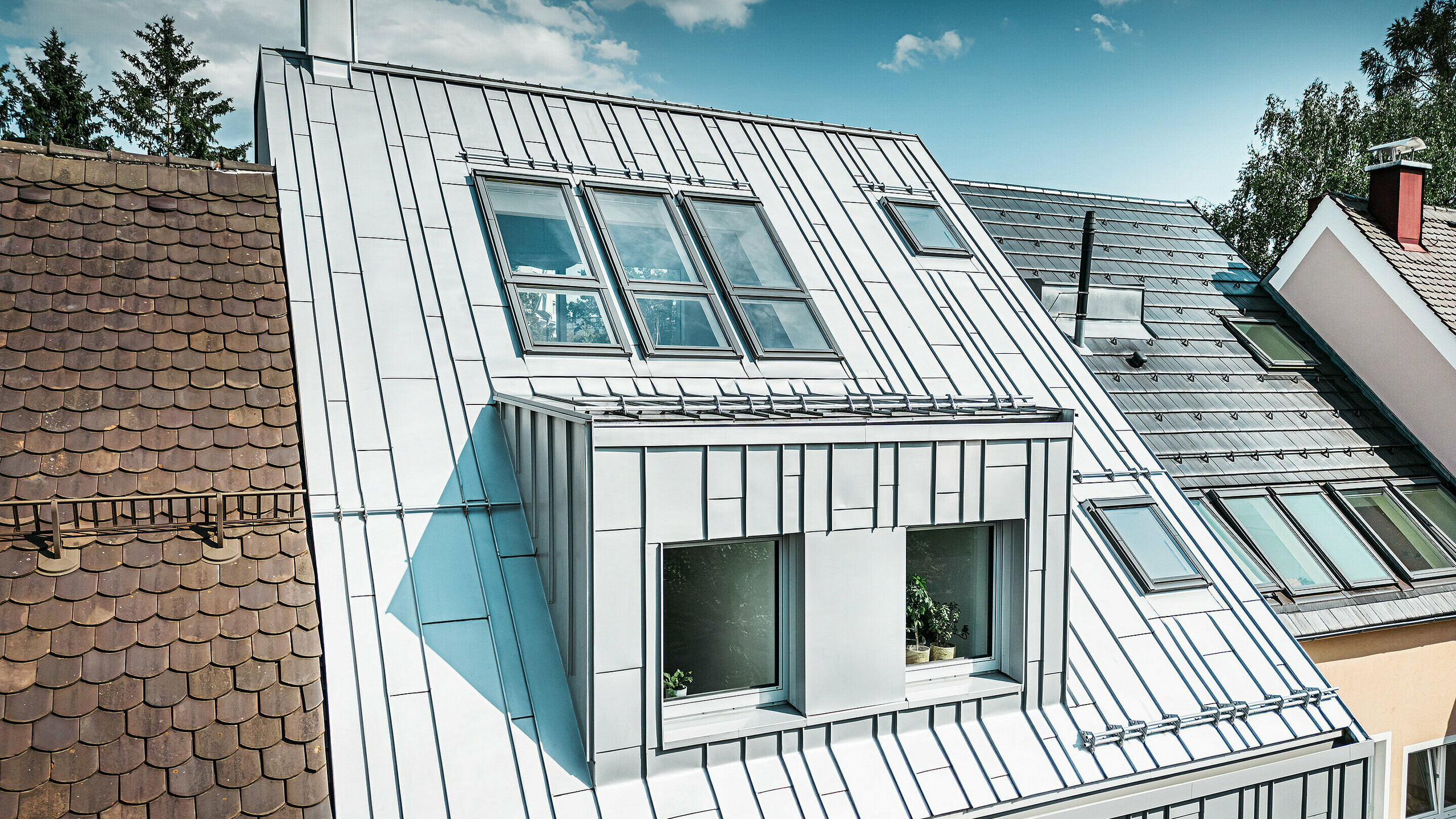 Une maison mitoyenne moderne dont le toit et la façade ont été rénovés, comme le montrent les produits en aluminium PREFA. Le toit est constitué de plaques d'aluminium argentées de petit format, à joint debout, qui présentent une surface structurée et mate et reflètent la lumière. La façade de la maison est également recouverte d'éléments verticaux en aluminium qui offrent un contraste contemporain avec les toits en tuiles traditionnels de couleur terracotta des maisons voisines.