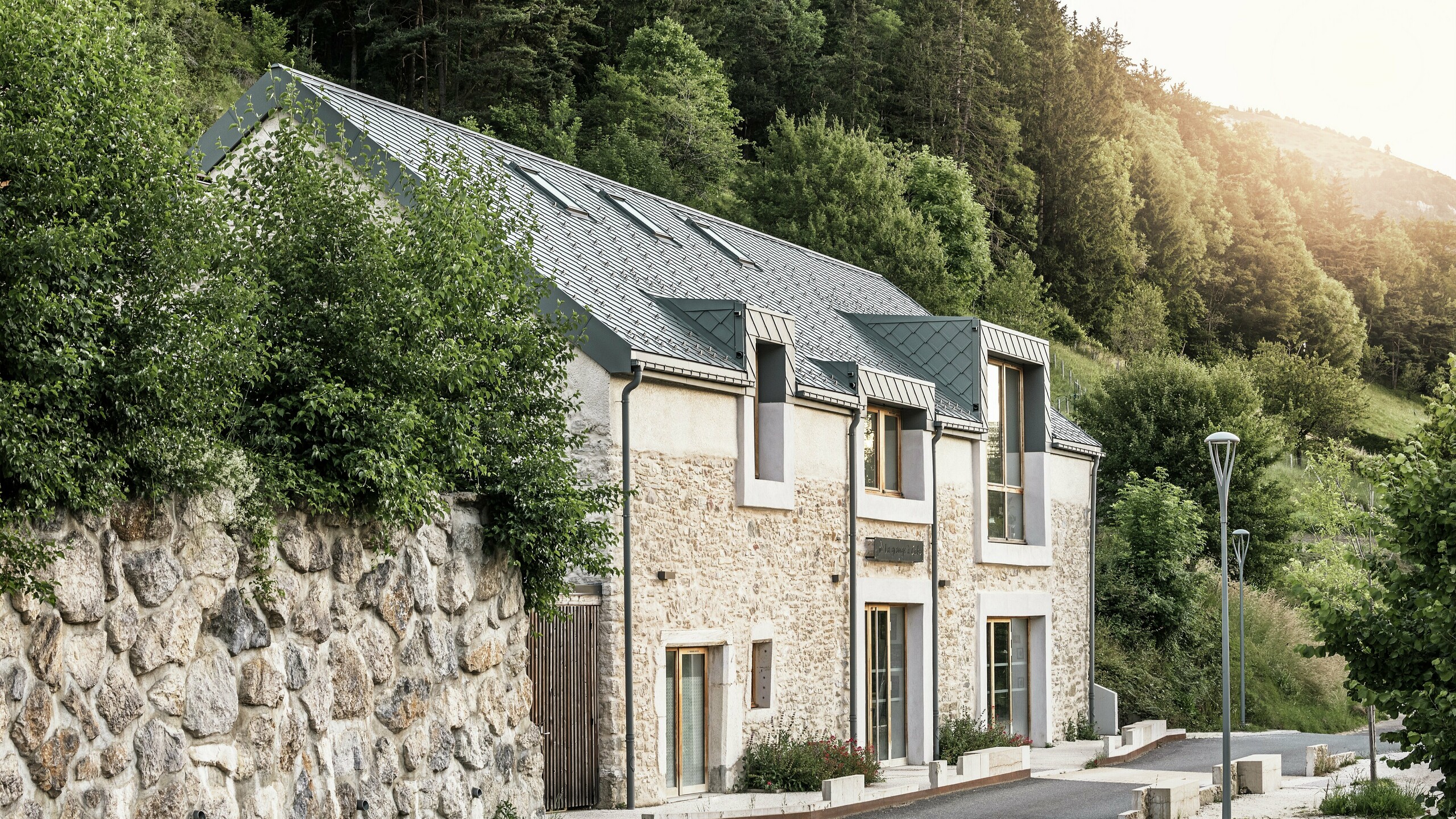 Vue latérale du corps de ferme rénové "La Grange à Gaby" à Engins, France. Le bâtiment combine la maçonnerie en pierre traditionnelle avec un toit moderne en aluminium PREFA composé de losanges de toiture de 29 × 29 cm. La rénovation soignée allie une construction historique à des matériaux durables.