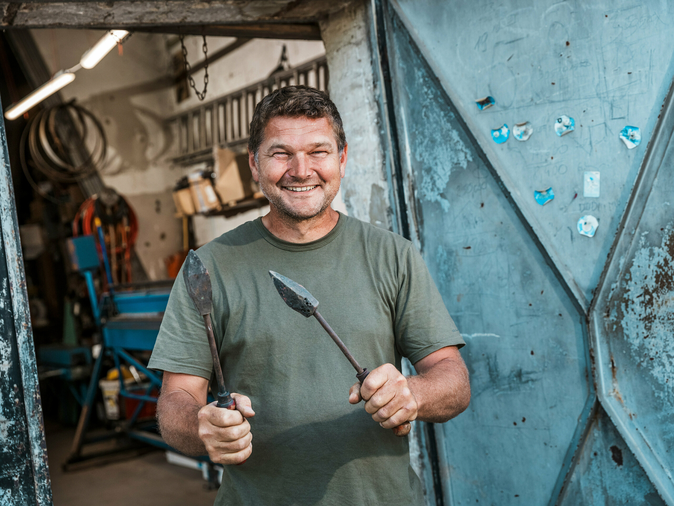 Le maître ferblantier Richárd Tóth dans son atelier. Cet artisan, qui se définit lui-même comme tel, tient des outils traditionnels devant une porte métallique ouverte. La scène témoigne d'un savoir-faire authentique et d'une grande précision, qualités essentielles à la fabrication des produits en aluminium PREFA pour l'immeuble d'appartements de Budapest. L'atmosphère brute de l'atelier souligne l'expertise artisanale avec laquelle les éléments de façade, le parapet et le revêtement de balcon ont été réalisés en PREFALZ, coloris P.10 anthracite. L'image met en valeur l'alliance de l'expérience, de la maîtrise des matériaux et de la durabilité des solutions PREFA.