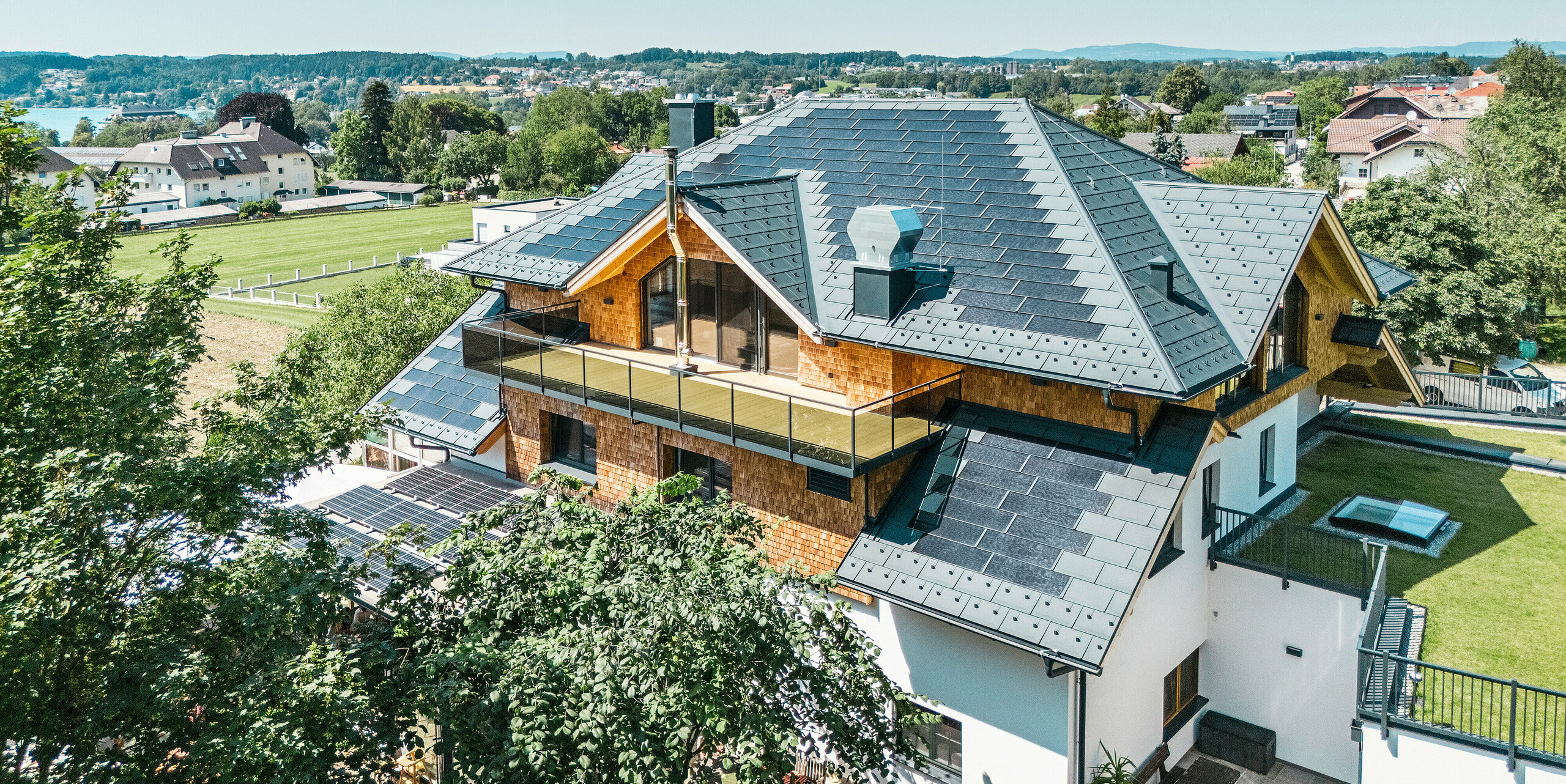 Vue aérienne du Zenz'n Stub'n à Schörfling am Attersee avec un toit récemment rénové en tuiles solaires PREFA et panneaux de toit R.16 en P.10 anthracite. La toiture en tôle de haute qualité allie un design moderne à une production d'énergie durable. Le balcon spacieux et la gouttière harmonieusement intégré soulignent l'aspect élégant du bâtiment.